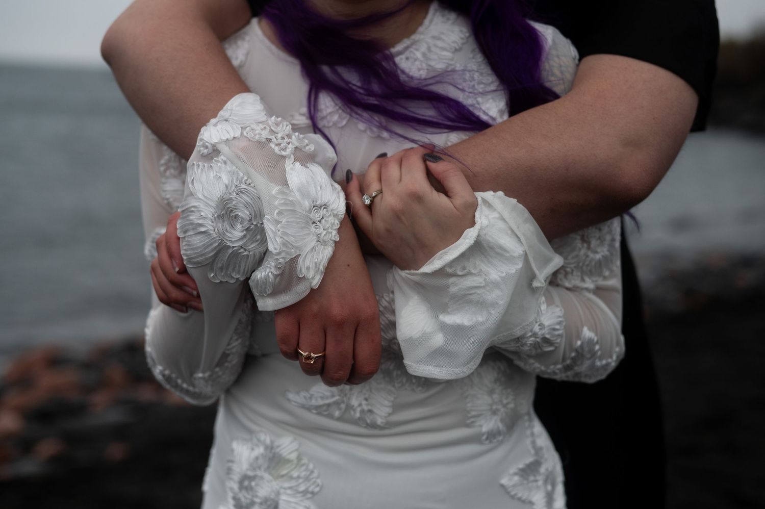 Bride in white lace wedding dress with purple hair embraced by groom near ocean shoreline.