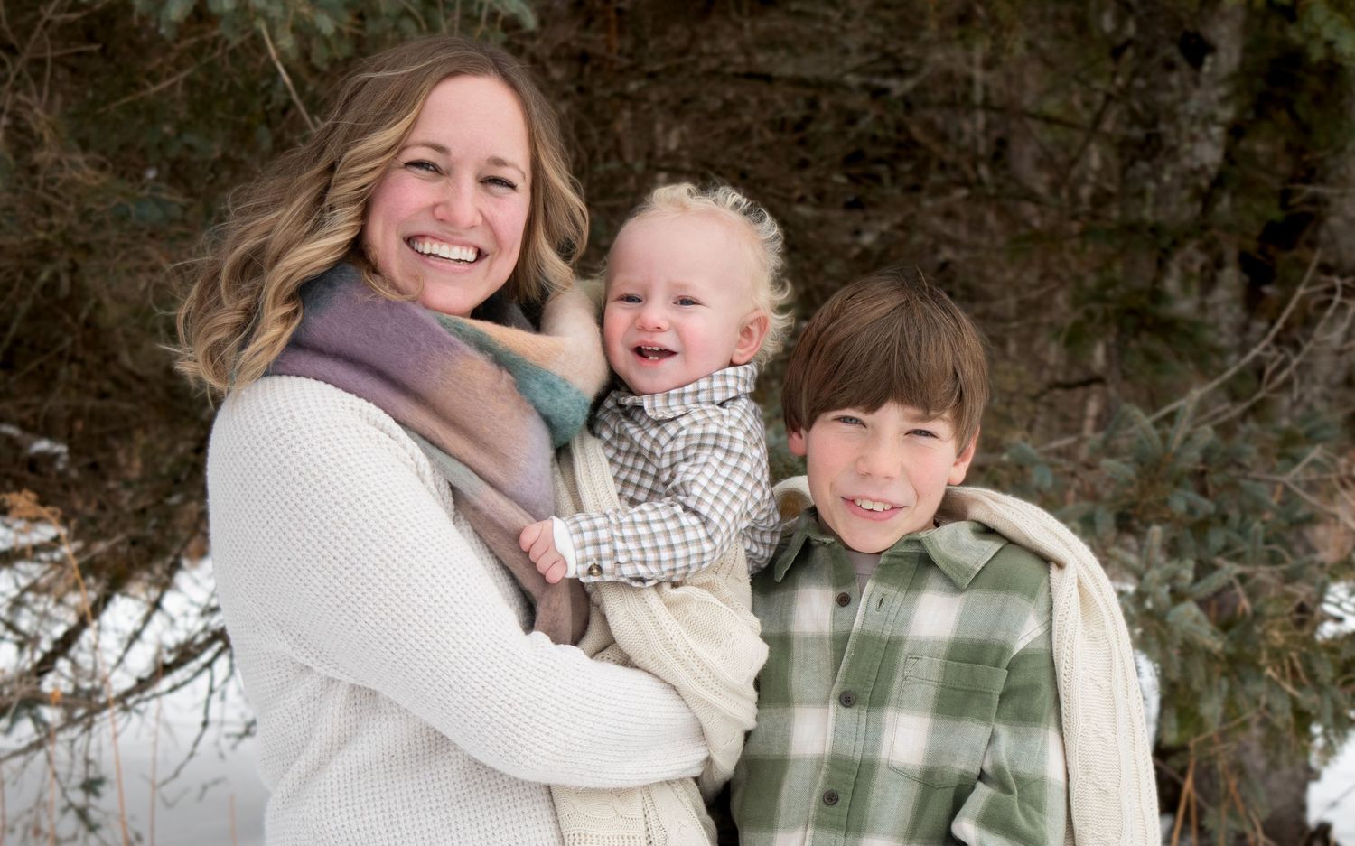 Mother in white sweater holding baby while standing with young son in snowy forest.