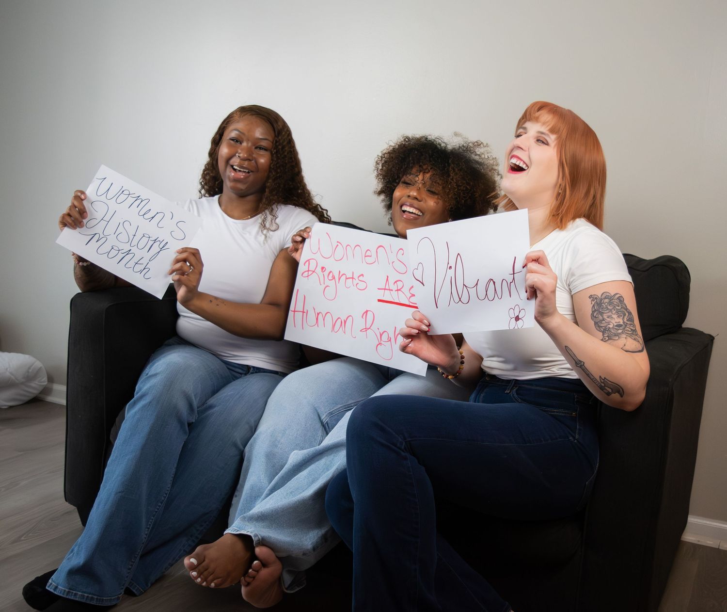 Three diverse women laughing and holding handwritten signs on a dark sofa.