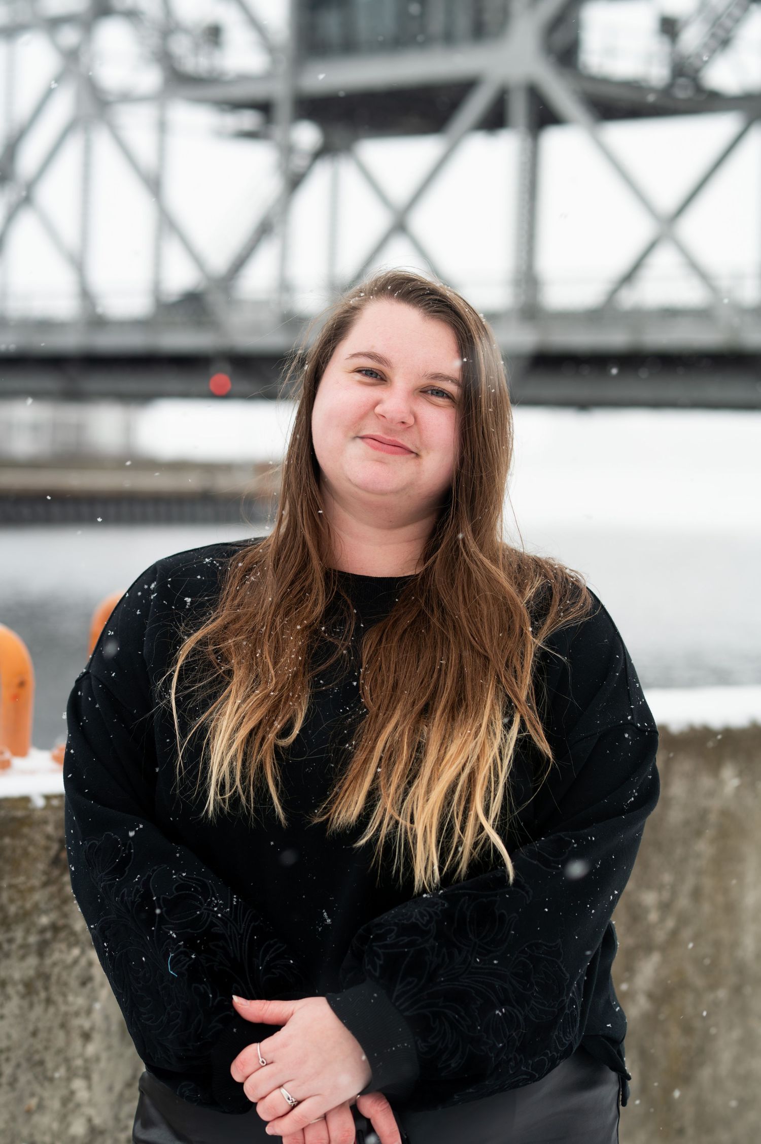 Young woman with long hair smiling outdoors near a bridge in winter, wearing a black sweater with light snow falling.