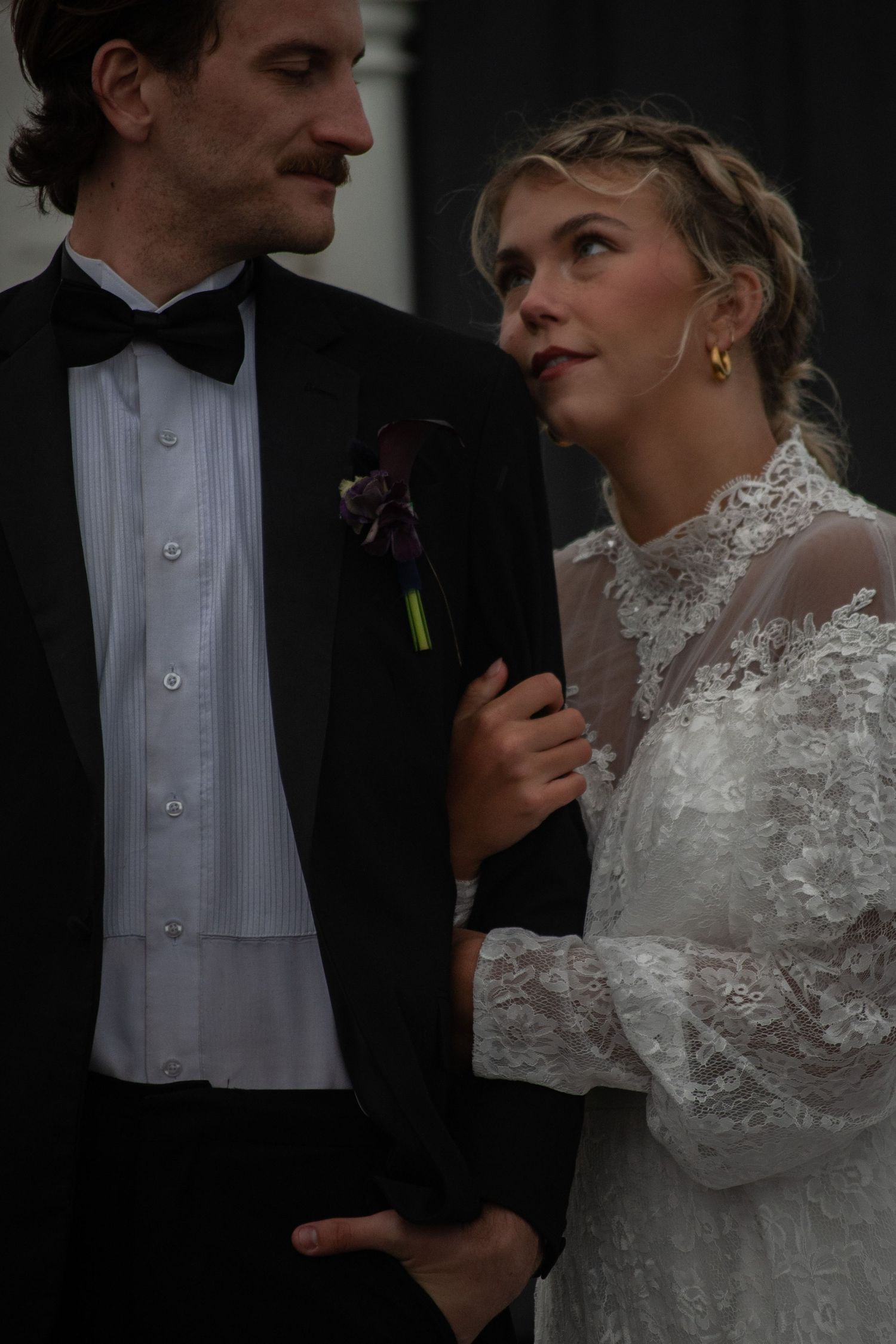 Bride in lace gown gazes at groom in black tuxedo with boutonniere during intimate wedding portrait.