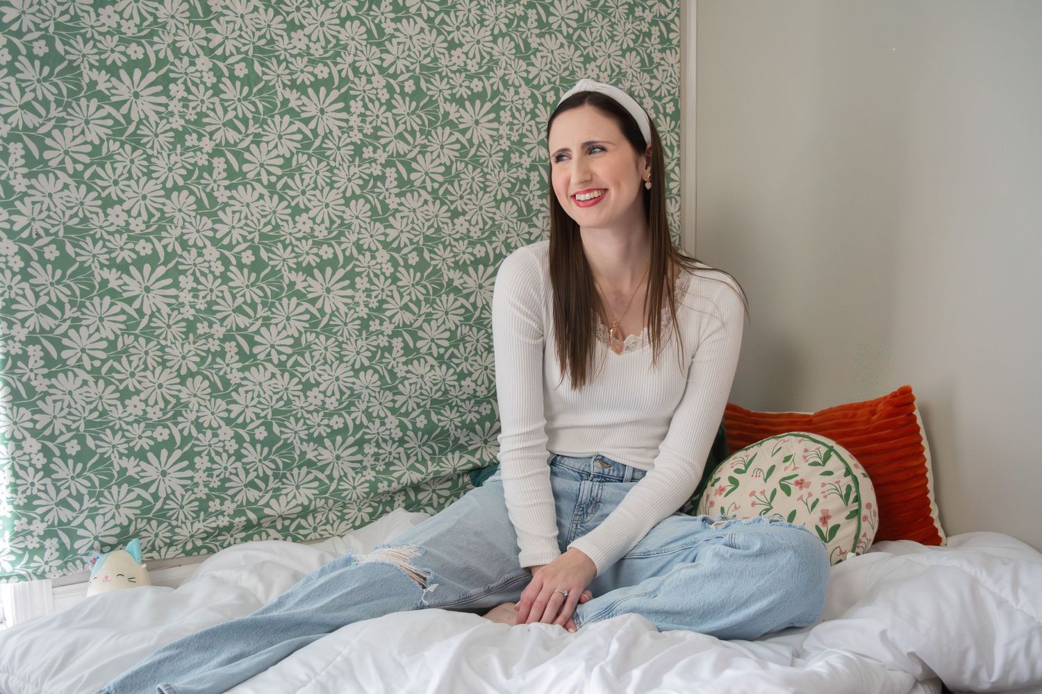 Smiling brunette woman sitting on a bed with green floral wallpaper backdrop.