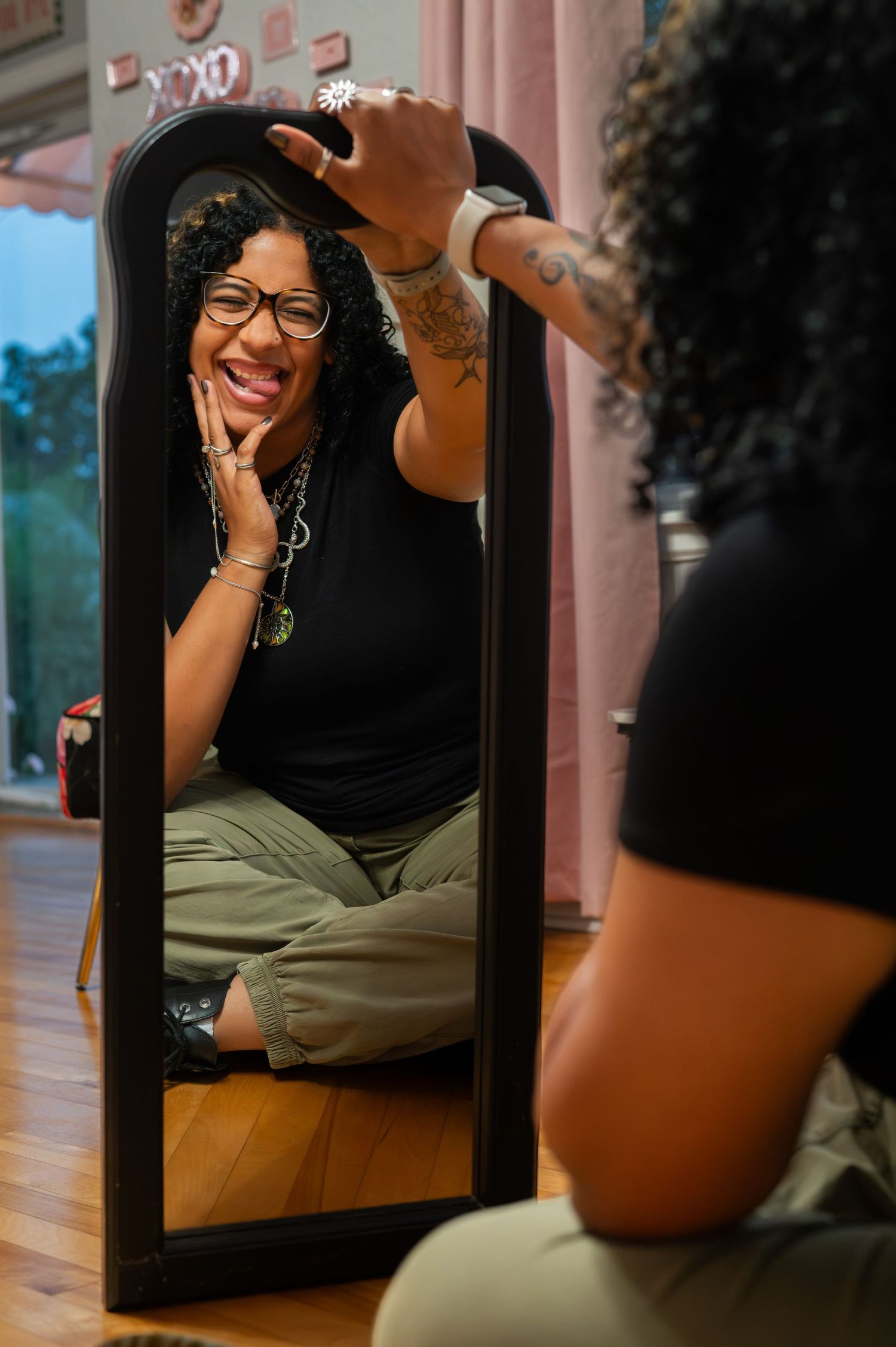 A smiling woman with glasses and tattoos sits cross-legged on a wood floor, gazing at her reflection in a black mirror.