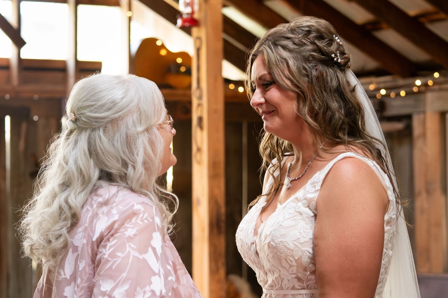 Emotional bride in lace wedding gown sharing a heartfelt moment with older woman in barn wedding venue.