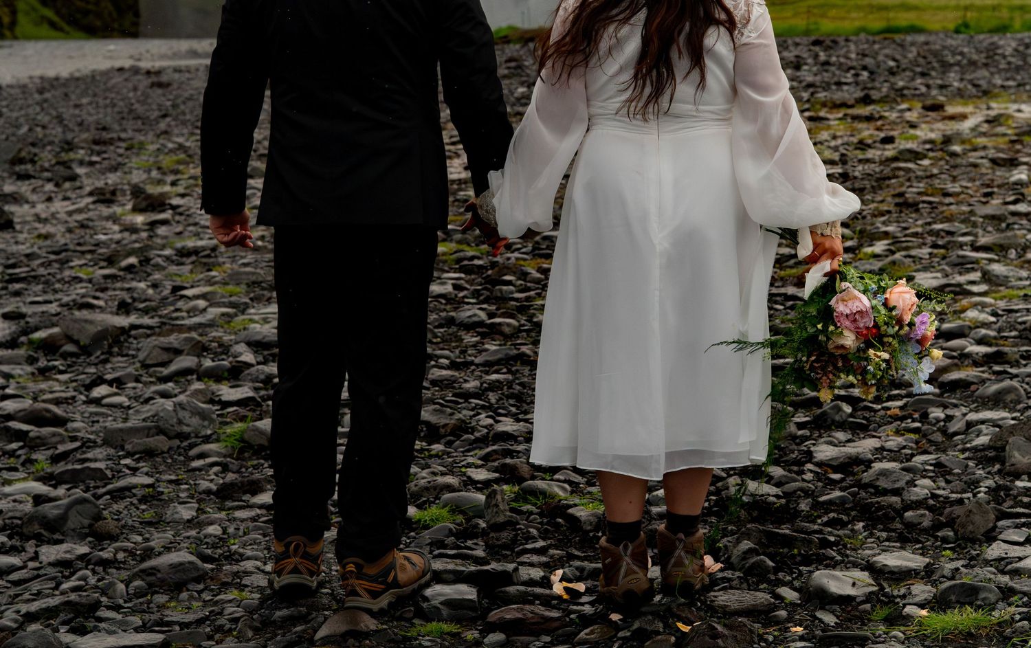 Bride and groom holding hands on rocky beach, woman in white dress holding bouquet, viewed from behind.