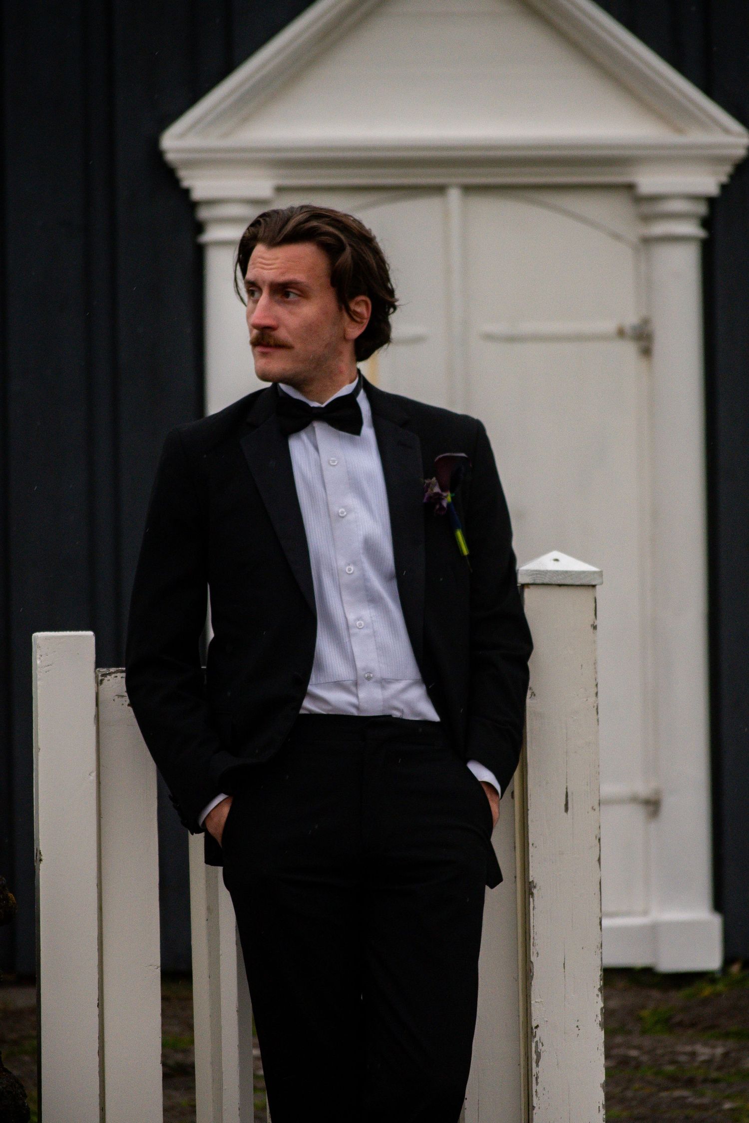 A groom in a black tuxedo with bow tie and boutonniere leans against a white fence outdoors.