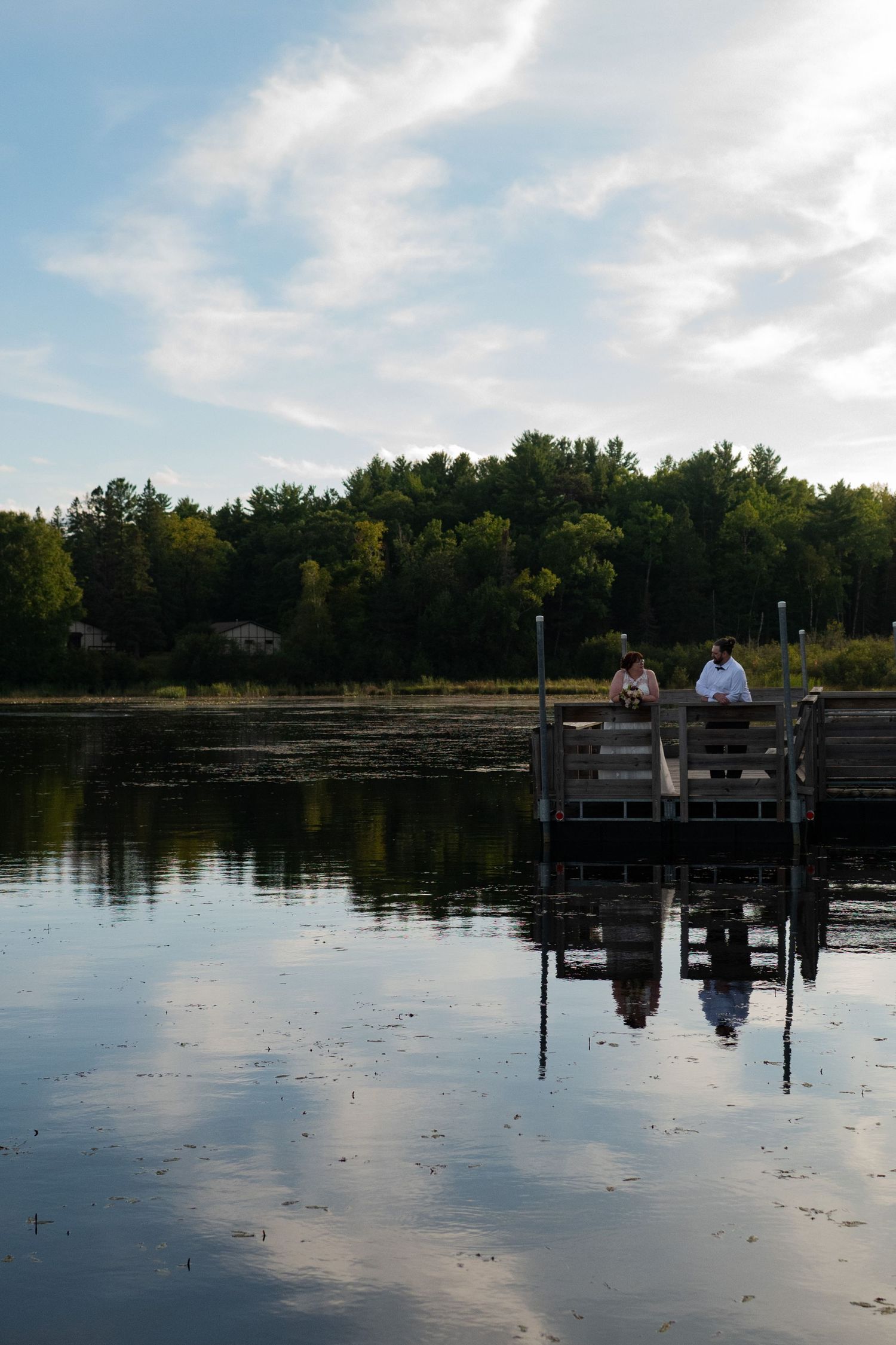 Two people stand on a wooden dock reflecting in a calm lake, surrounded by lush green trees under a partly cloudy sky.