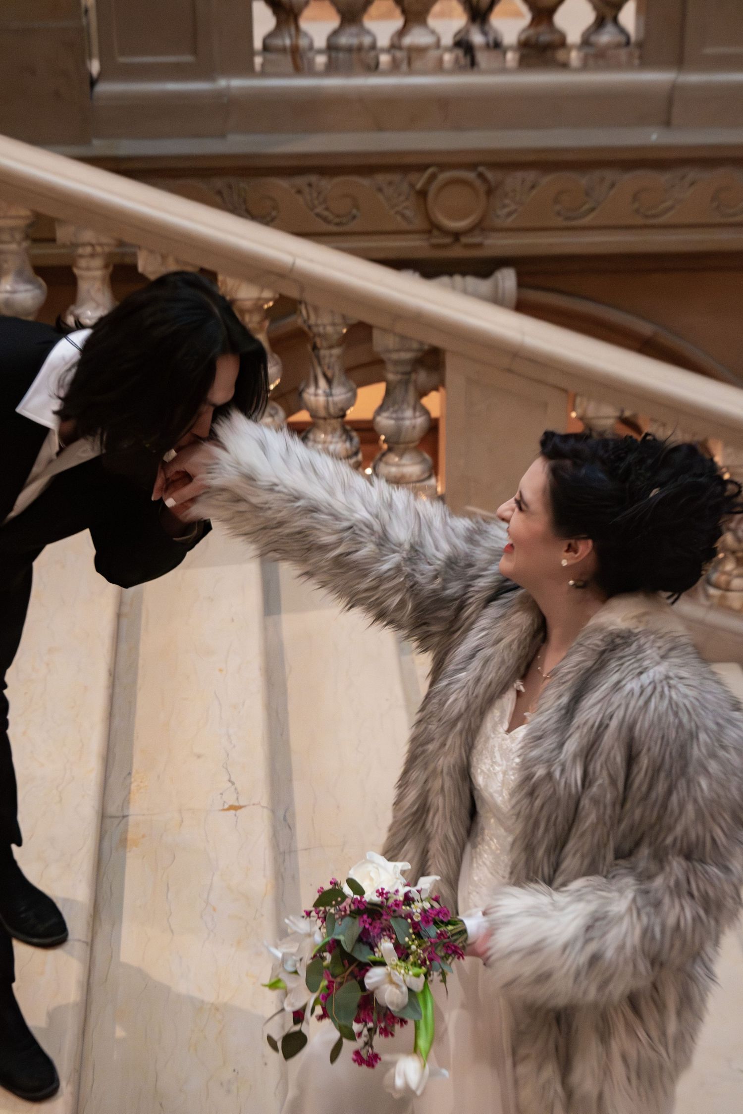 Bride in white dress and fur coat holds partner's hand on elegant marble staircase during wedding photos.