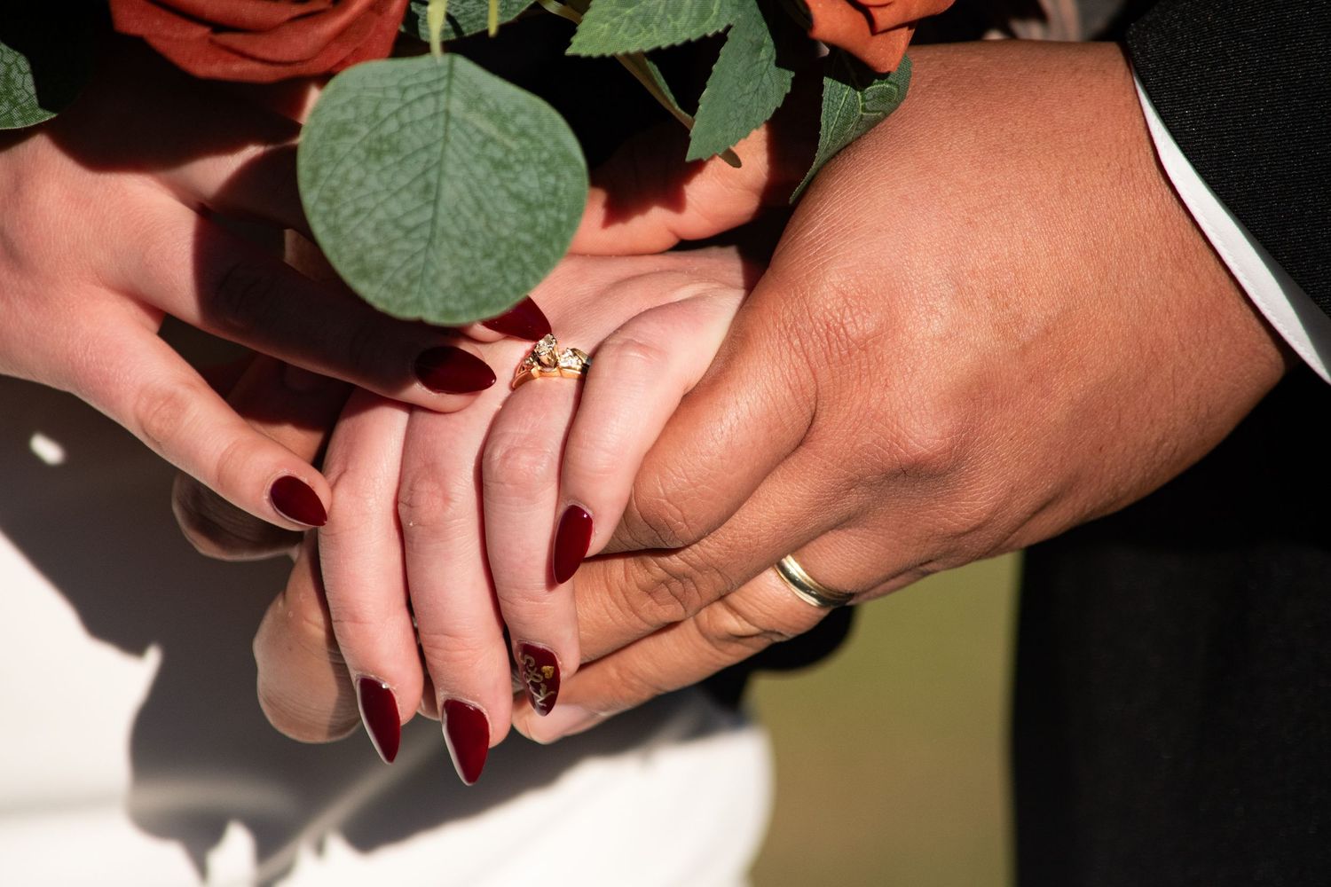 Bride and groom holding hands showing wedding rings with orange rose bouquet in background.