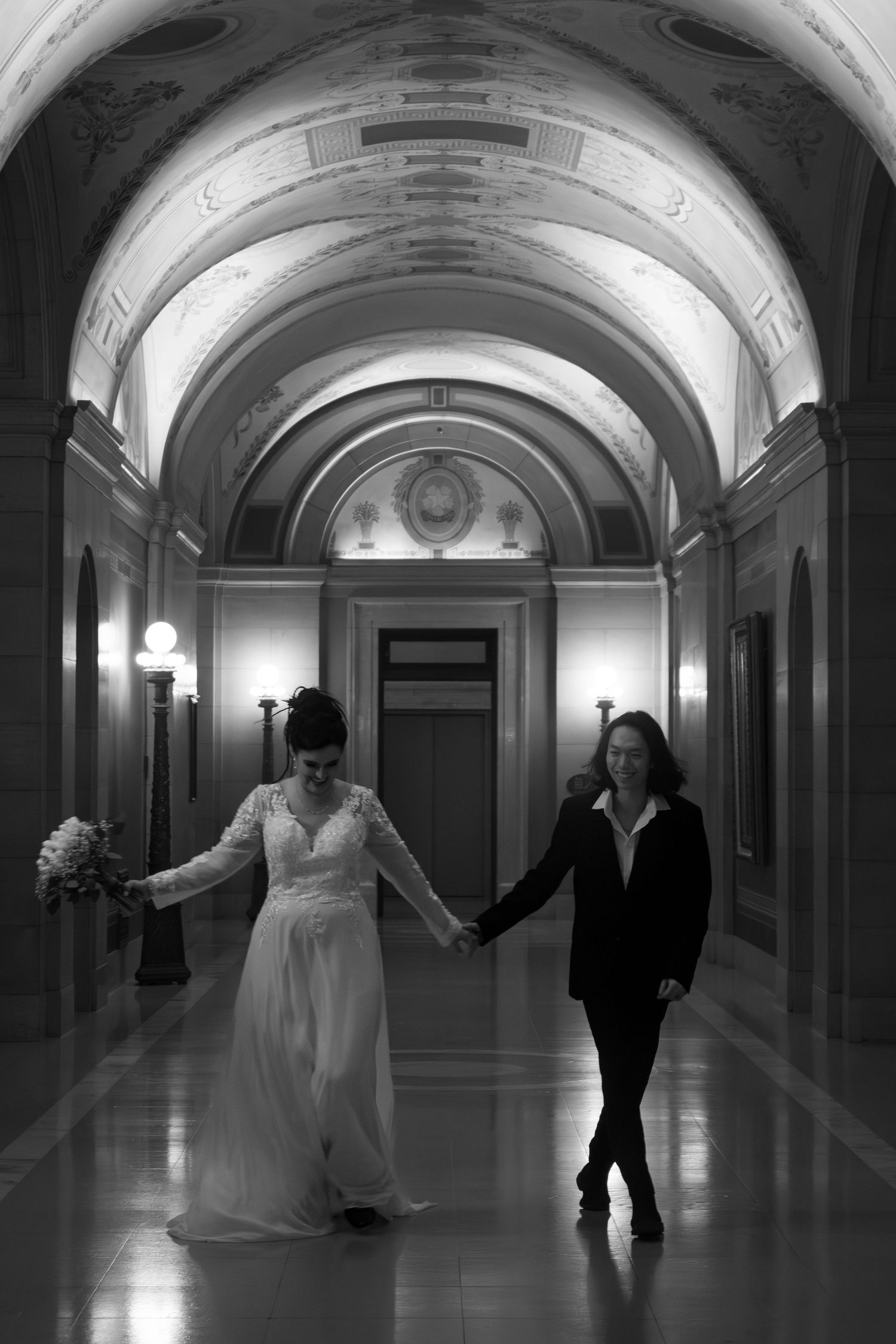 Black and white photo of two brides holding hands walking through grand arched hallway in historic building.