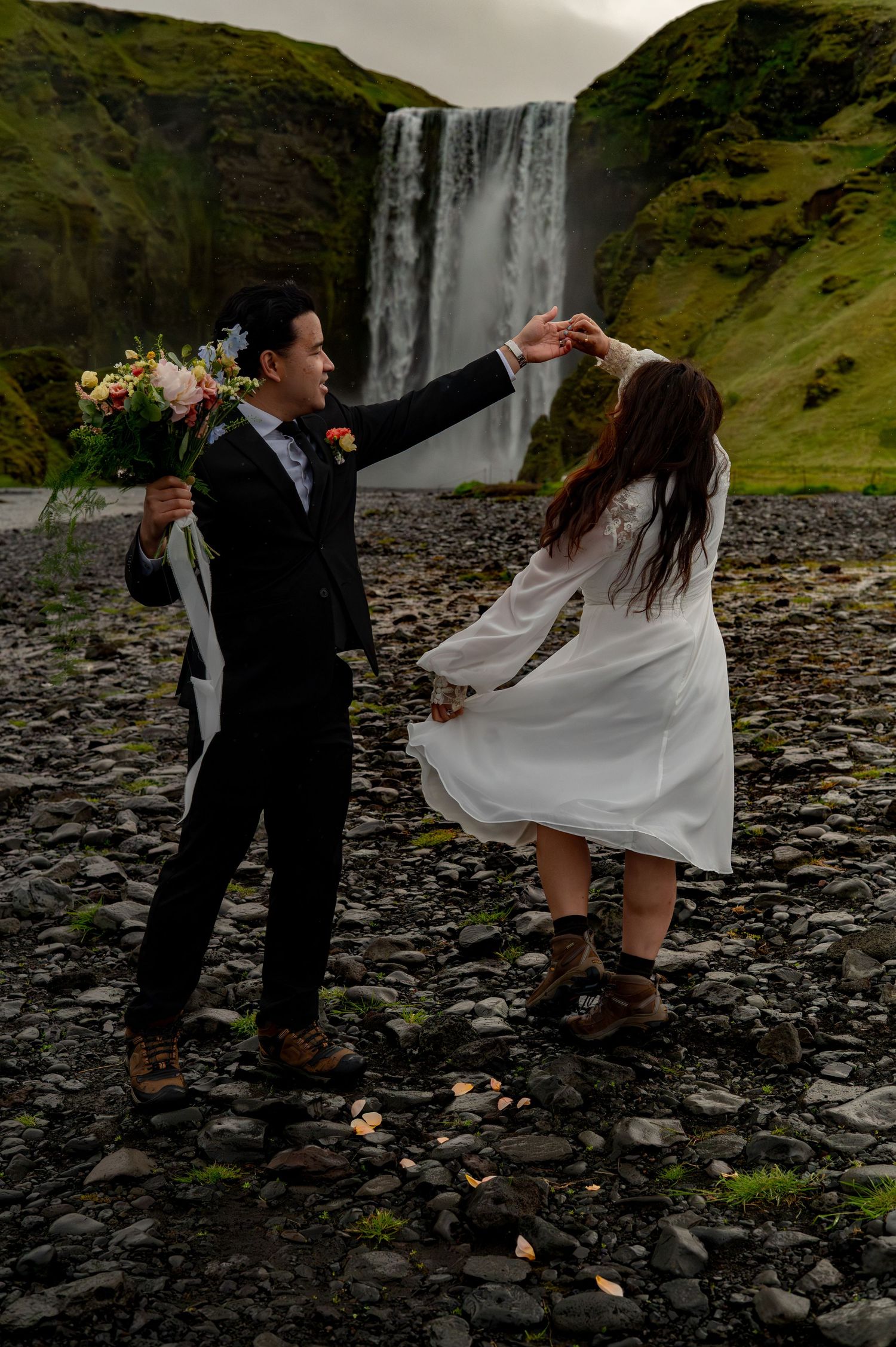 Couple dancing near Skogafoss waterfall in Iceland, groom in black suit holding bouquet, bride in white dress twirling.