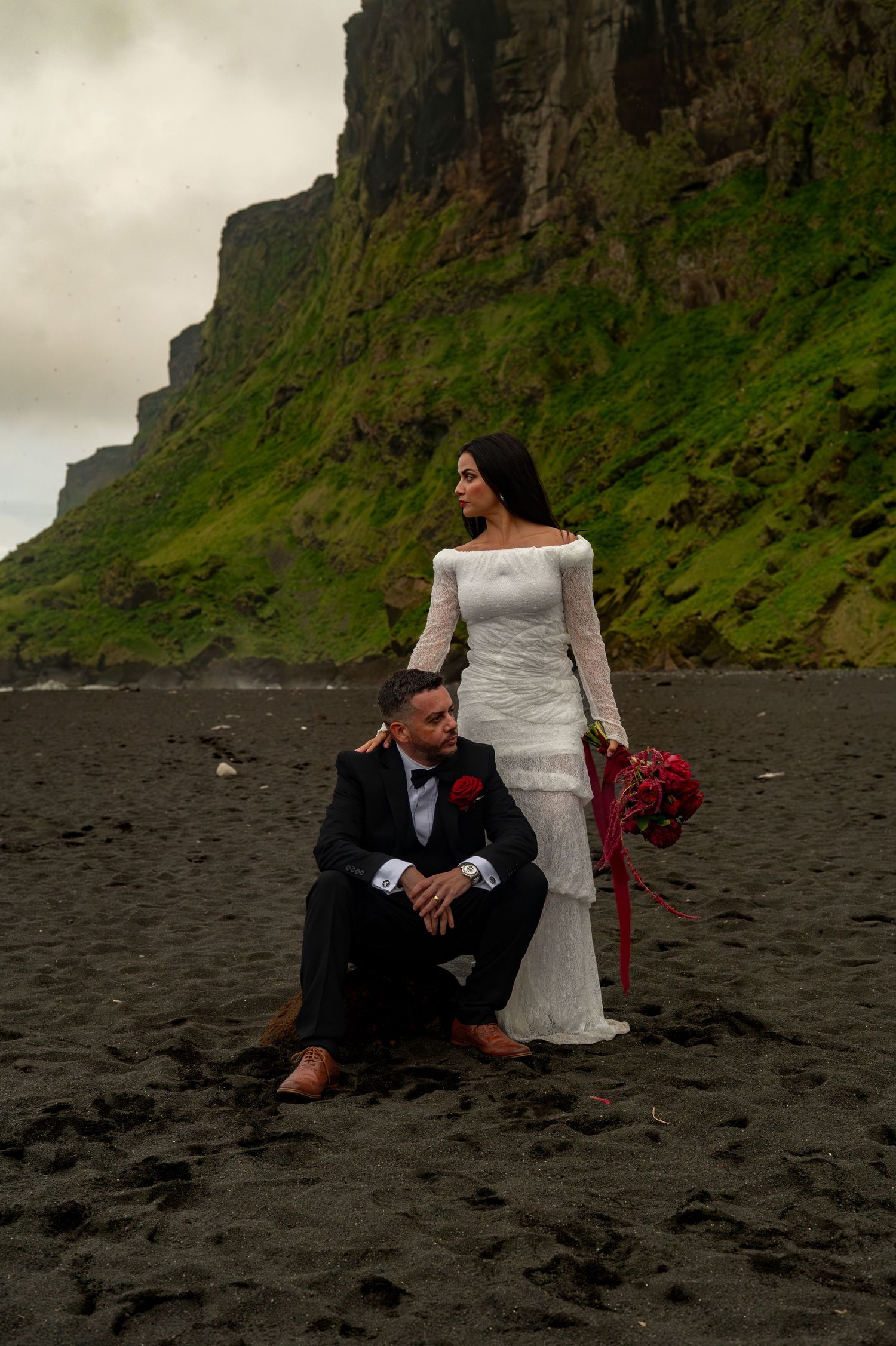 Bride and groom posing on black sand beach with dramatic green cliffs, bride in white gown holding red bouquet.