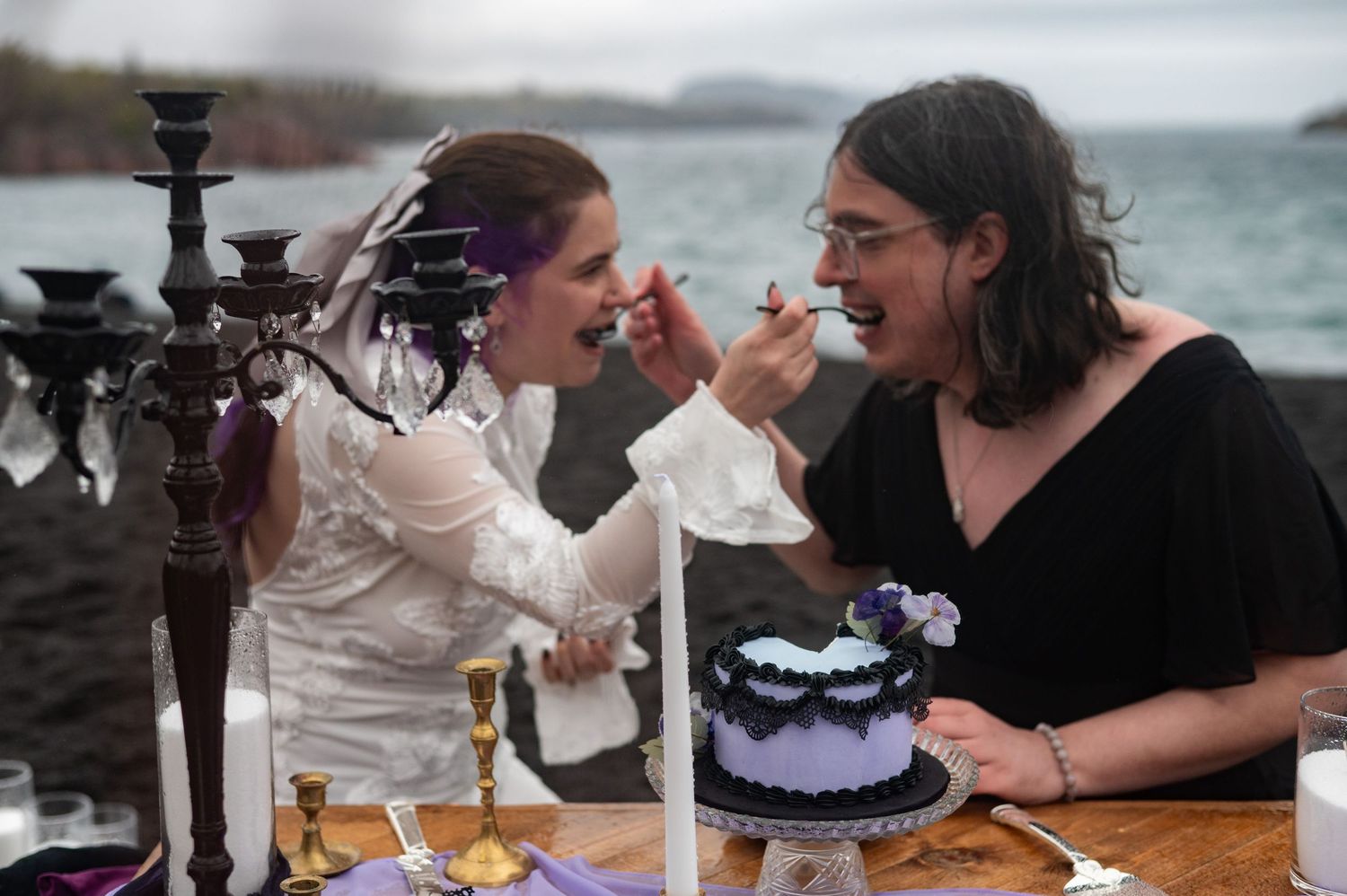 Couple sharing gothic wedding cake outdoors by rocky ocean shore, bride in white feeding groom at decorated table.