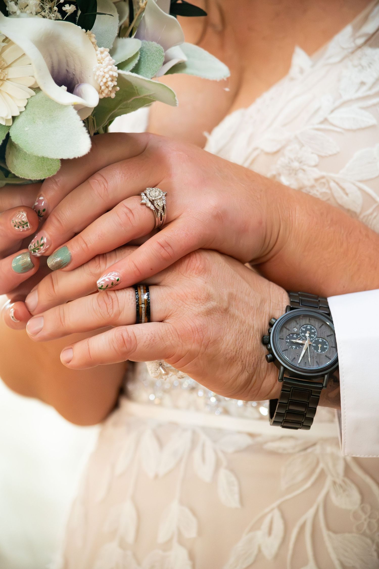 Bride and groom holding hands showing wedding rings and a black chronograph watch with a floral bouquet.