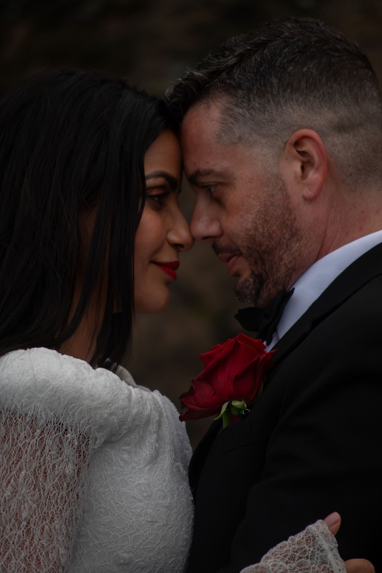 A bride and groom share an intimate moment, touching foreheads, as she wears white lace and he holds a red rose.