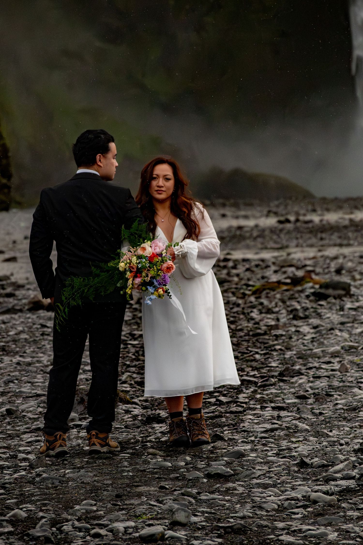 Couple in wedding attire stands on rocky shore near misty waterfall, bride holding colorful bouquet in white dress and boots.