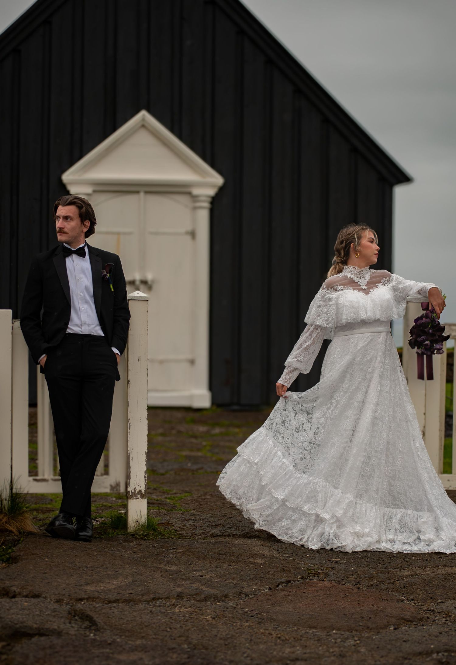 Bride in vintage lace gown and groom in tuxedo pose near black barn at outdoor wedding venue.