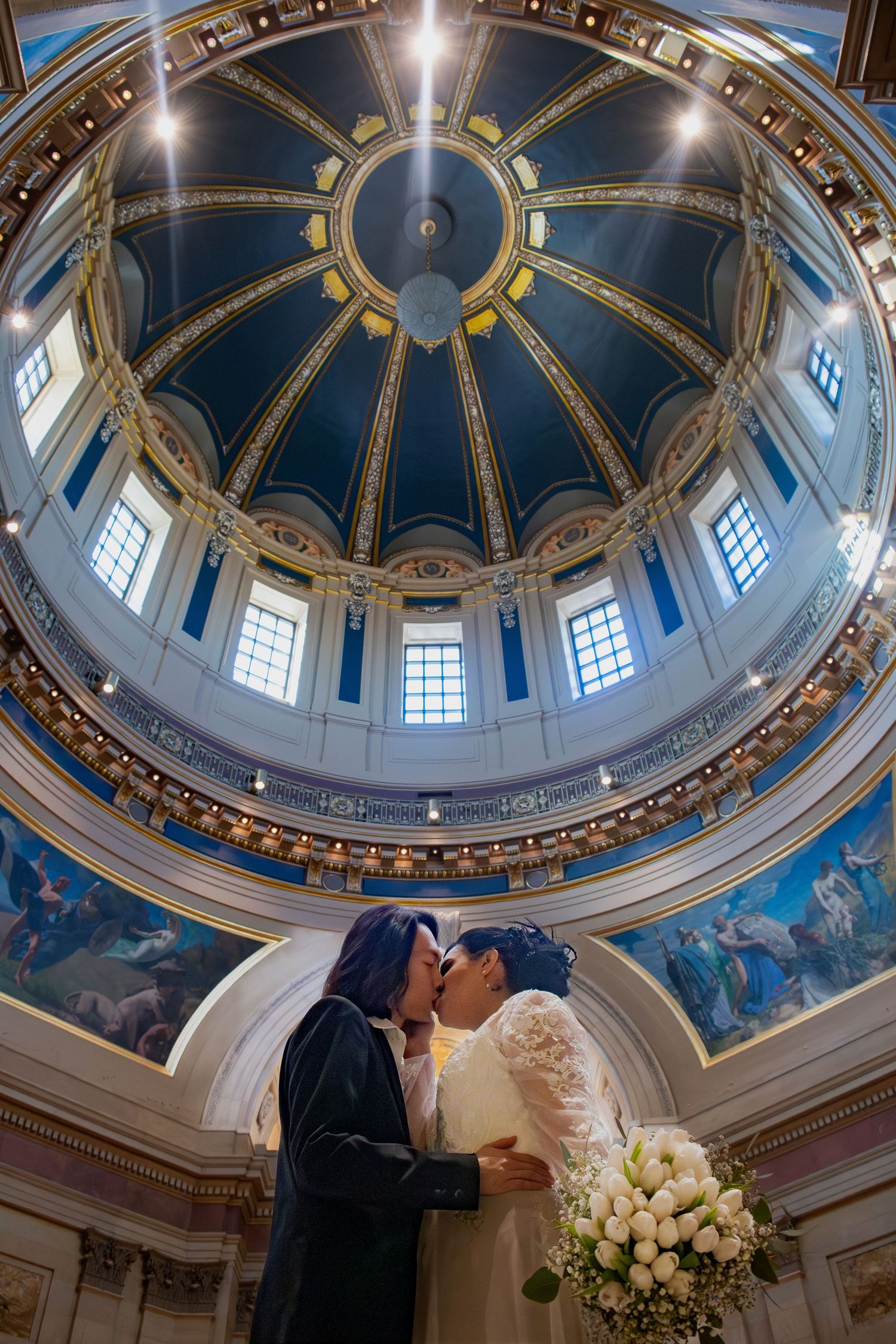 Newlywed couple shares first kiss beneath ornate blue and gold dome of a historic capitol building.