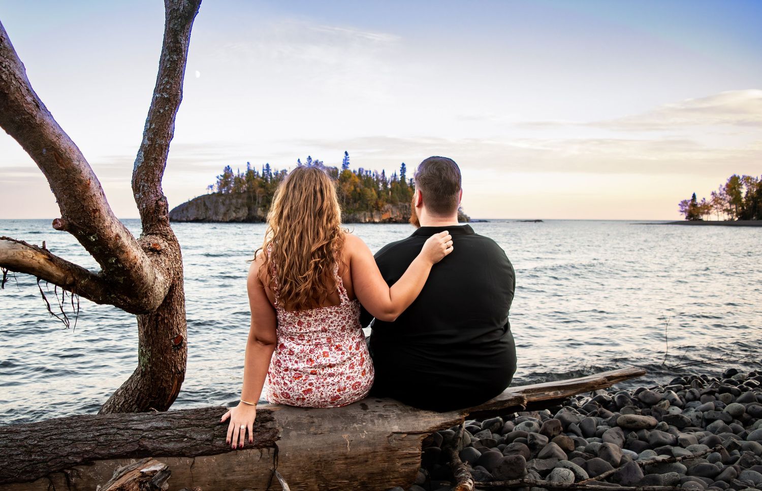 Couple sits on driftwood log embracing while overlooking a calm lake at dusk.
