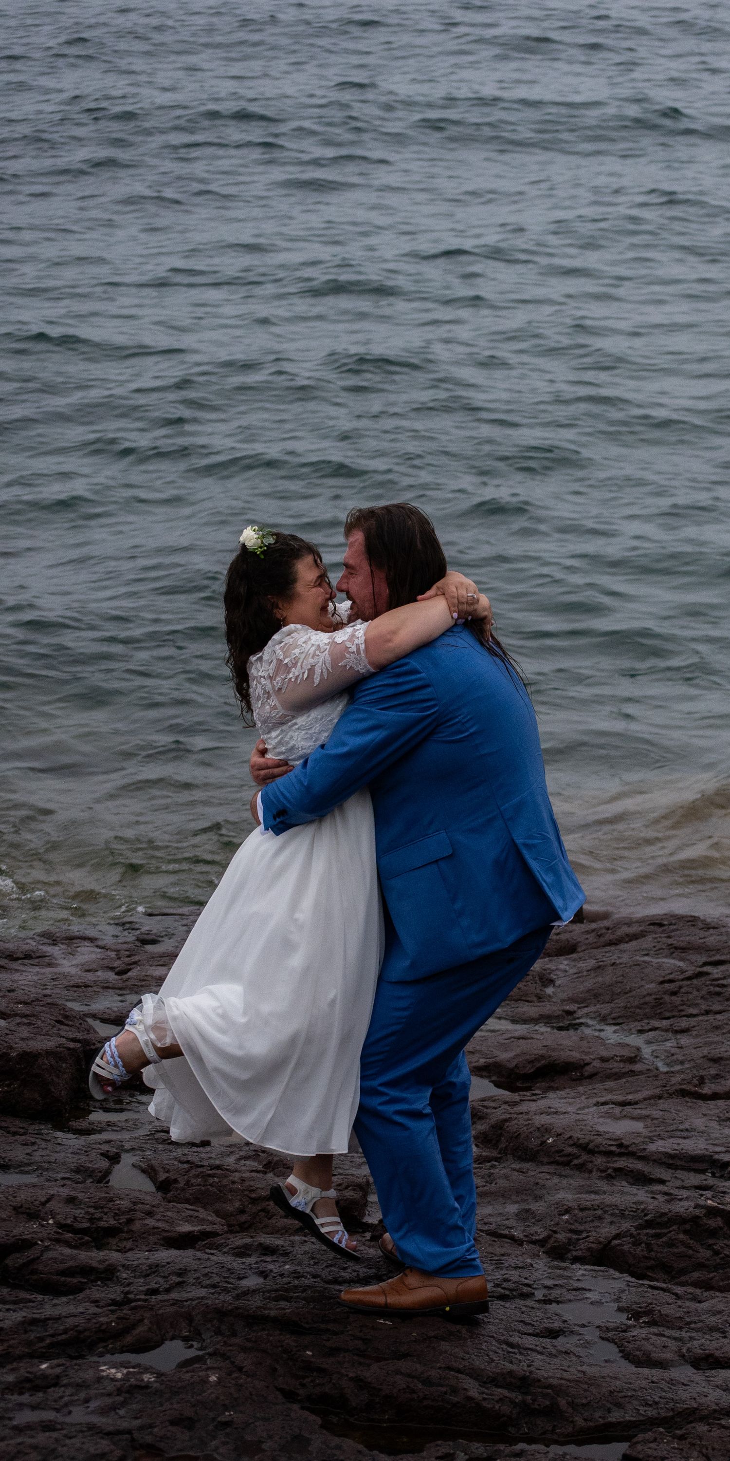 Groom in blue suit lifts bride in white lace dress on rocky shoreline with ocean waves behind them.
