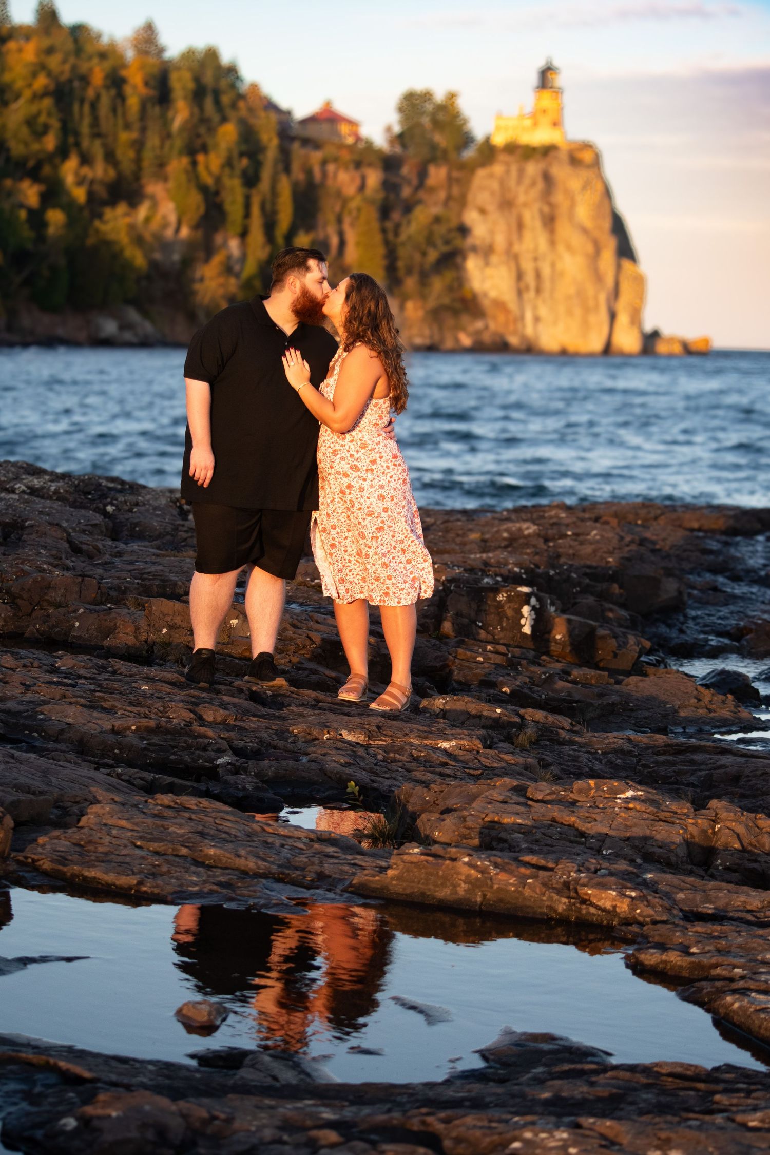 Couple sharing a kiss on rocky shoreline with Split Rock Lighthouse reflected in water below.