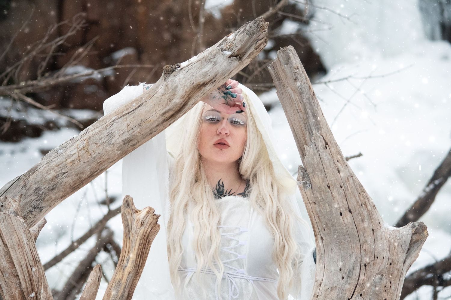 Tattooed blonde woman in white hooded gown framed by driftwood branches in snow.