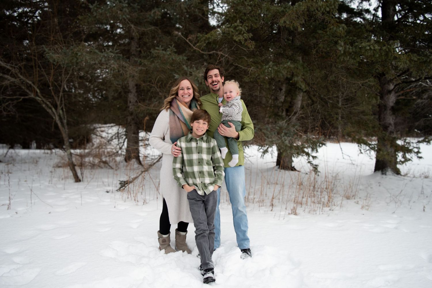 Smiling family of four standing together in snow-covered forest during winter family photo session.