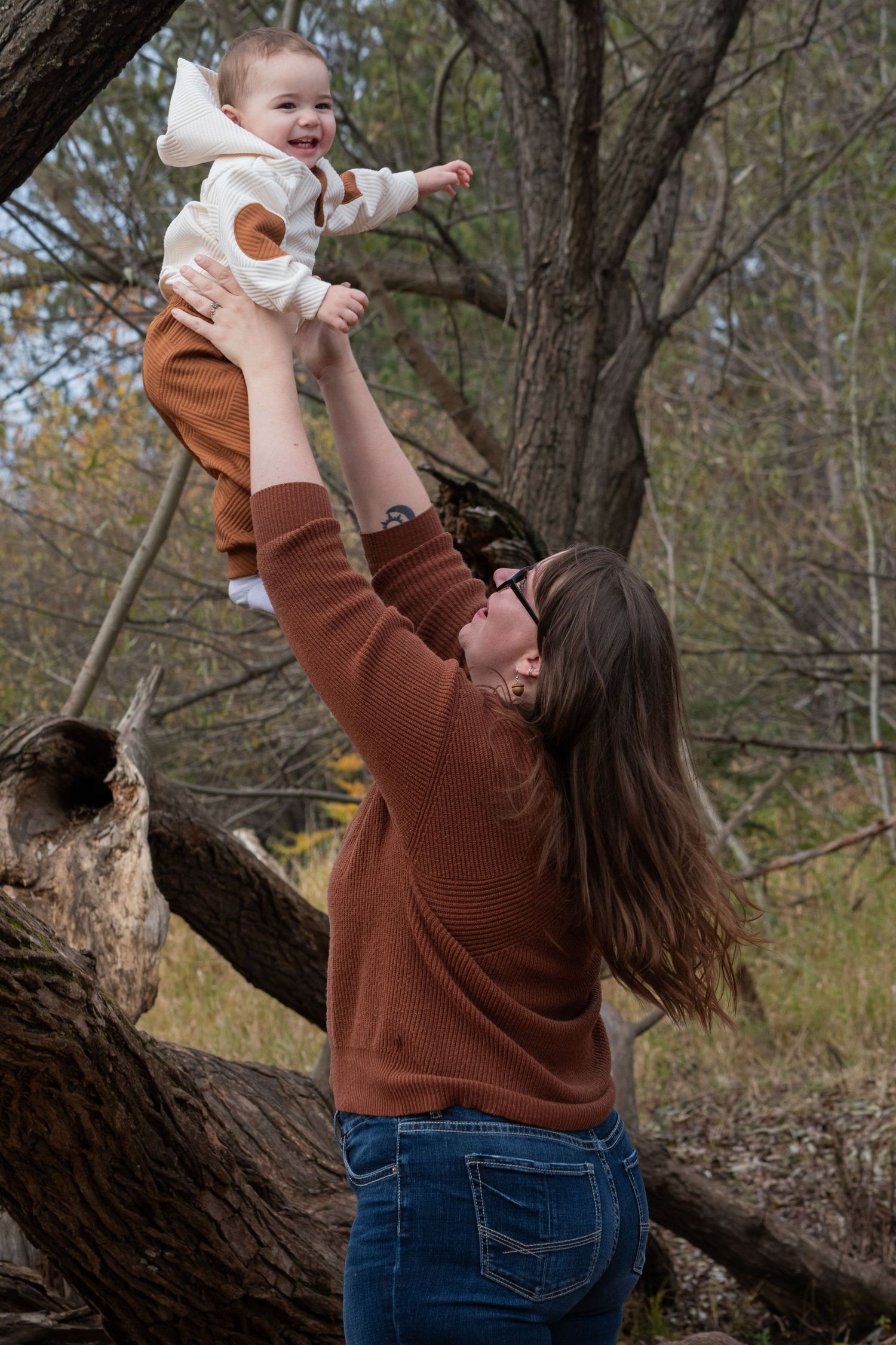 Mother in rust sweater lifting laughing baby overhead near trees in autumn.
