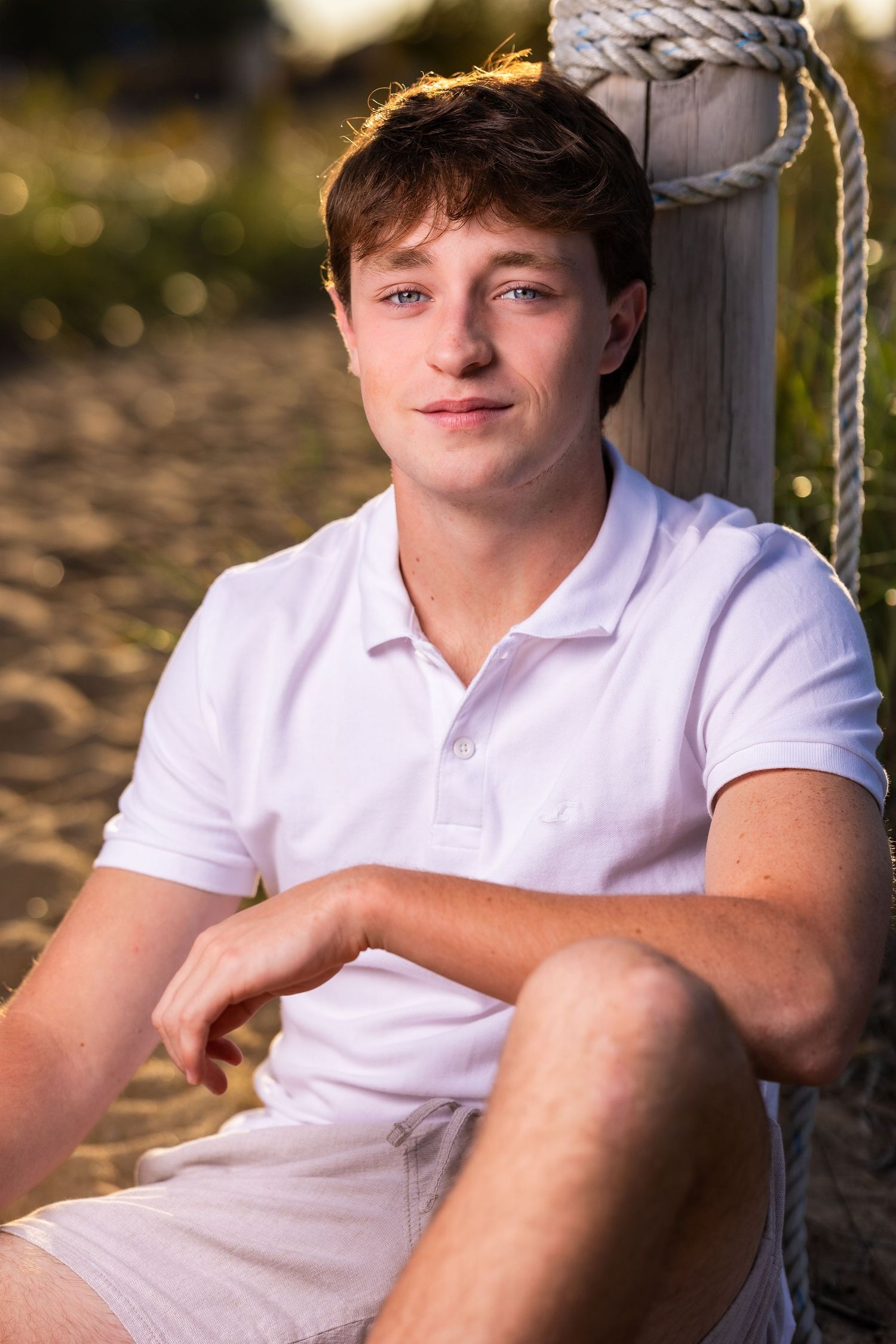 Person in white polo shirt sitting outdoors near wooden post at sunset.
