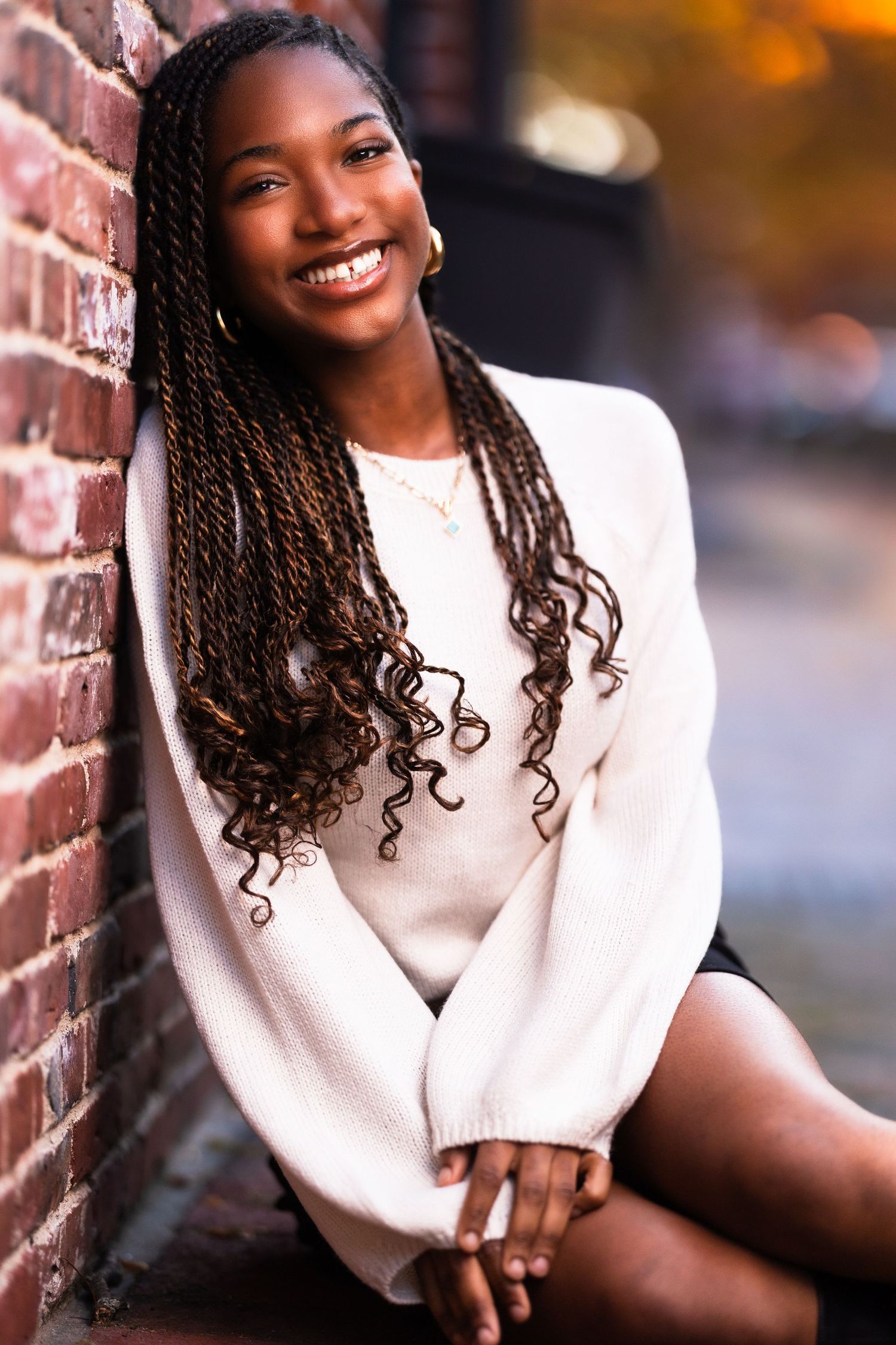 Person in white sweater poses against brick wall with warm lighting.