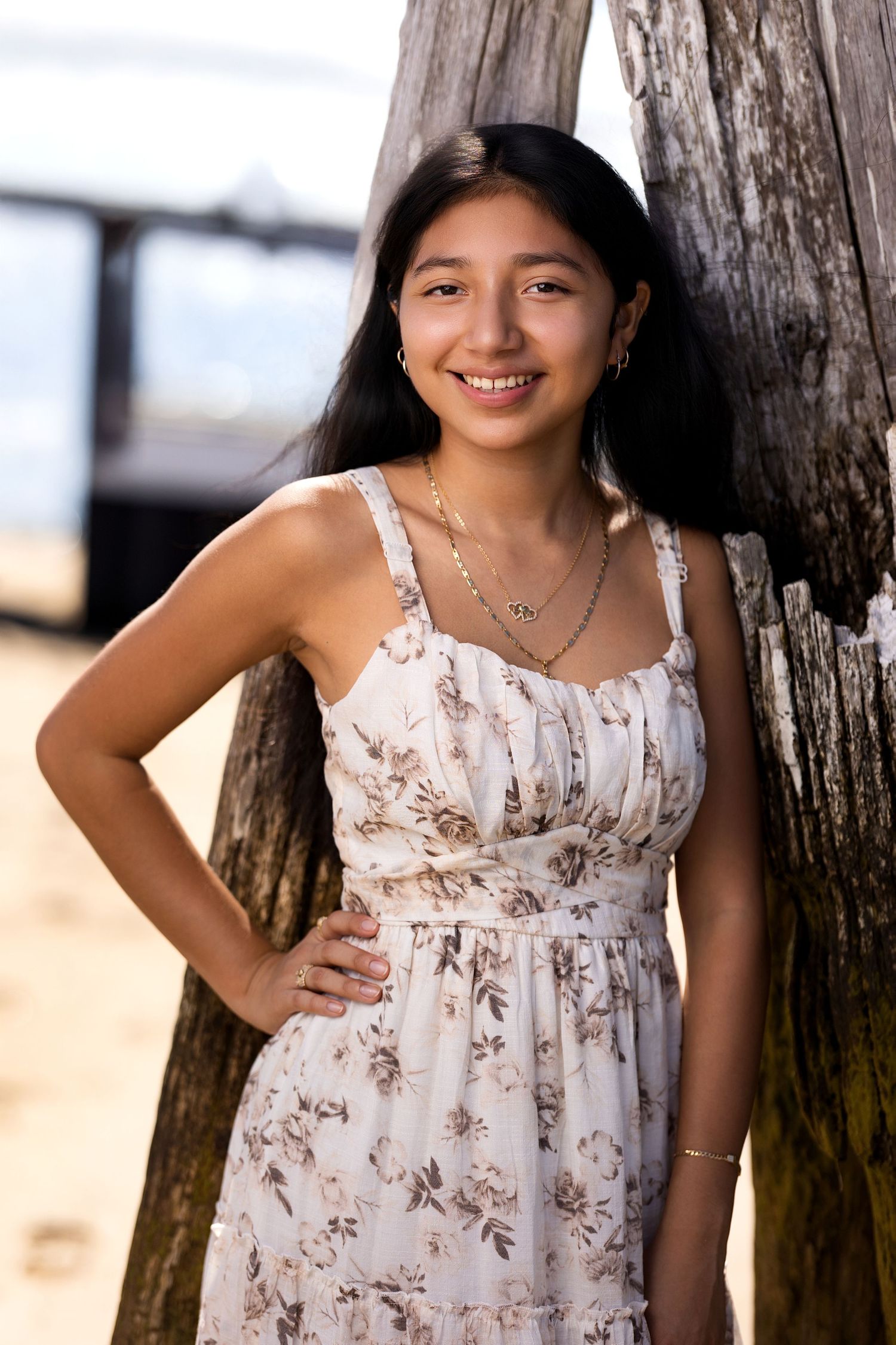 Person in floral sundress poses against wooden post at beach.