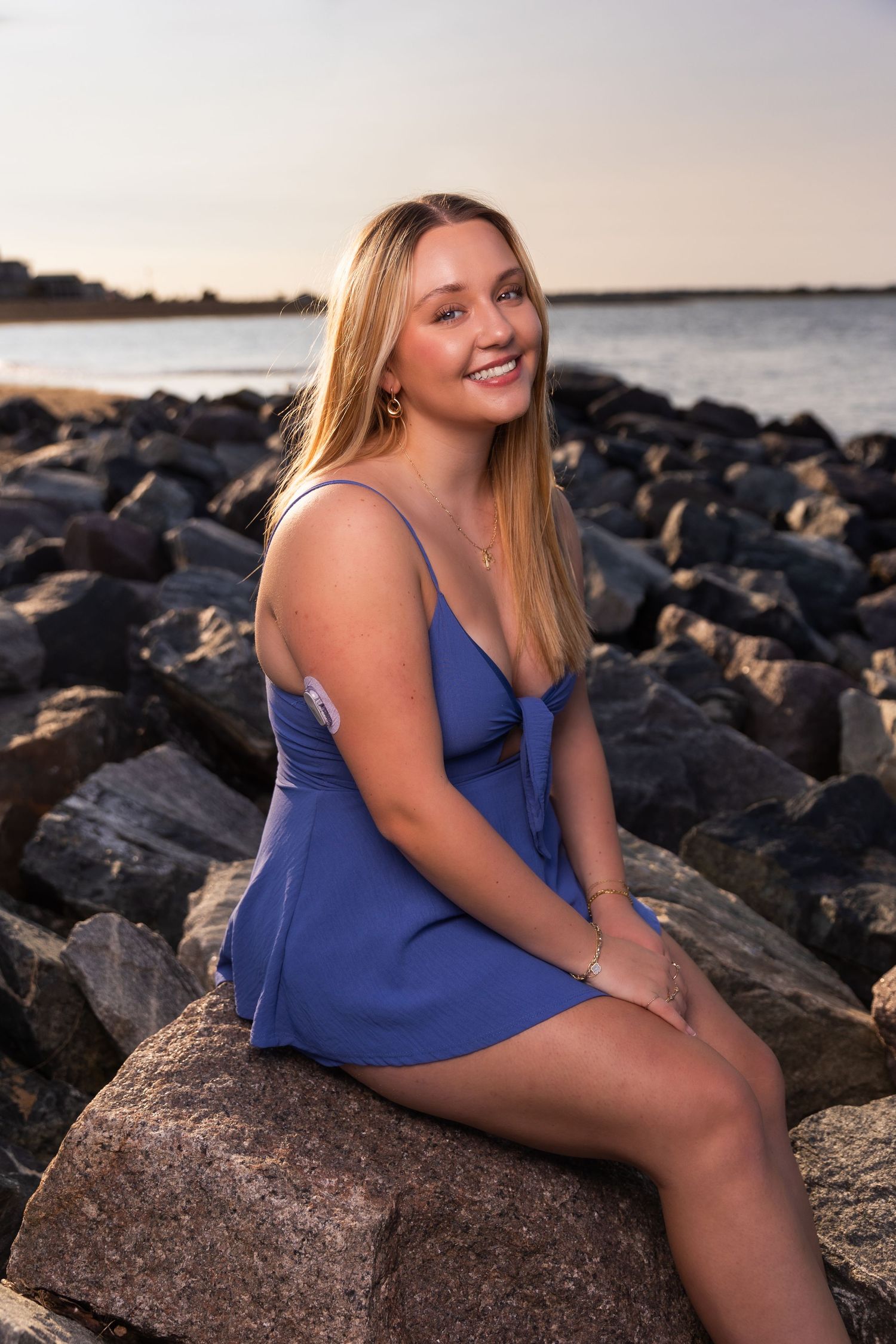 A person in a blue dress poses on rocky coastline during sunset.