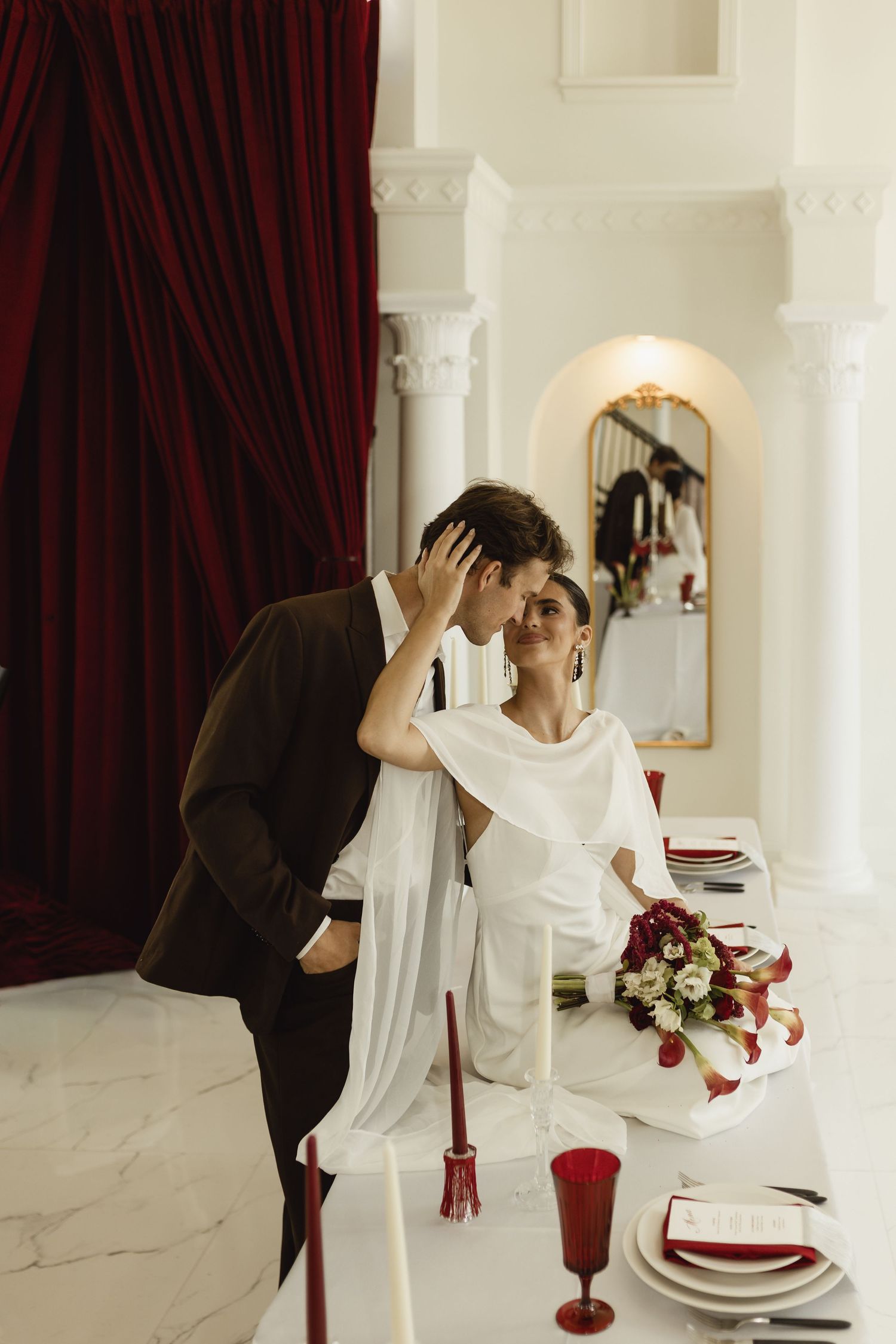 Elegant bride and groom share romantic moment at decorated wedding table with red accents and white marble interior.