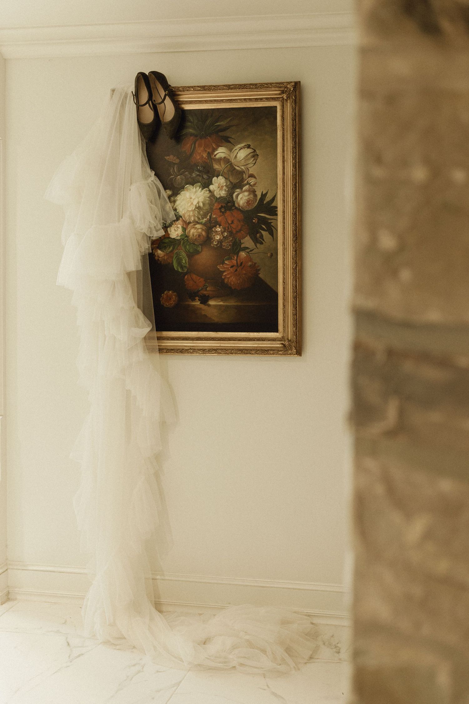 Bridal veil and shoes displayed against a floral oil painting in a gold frame on a marble floor.
