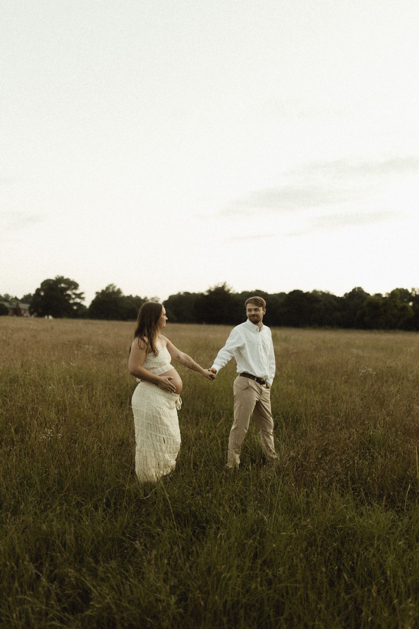 Expecting couple holds hands walking through golden meadow at sunset, woman in white lace maternity dress.