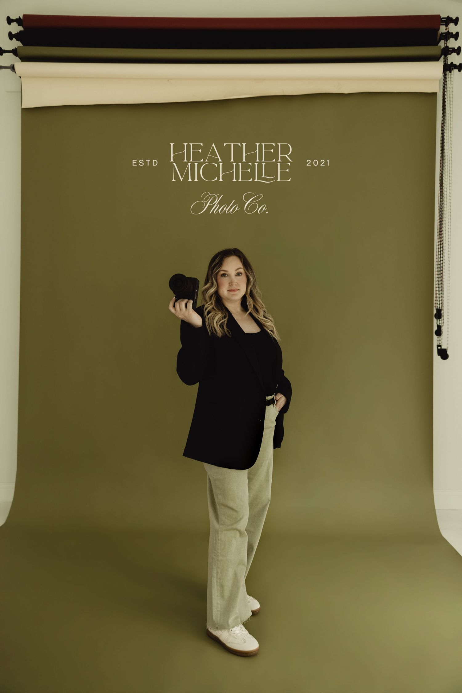 Photographer holding camera in studio with olive green backdrop, wearing black top and tan pants, Heather Michelle Photo Co. logo.