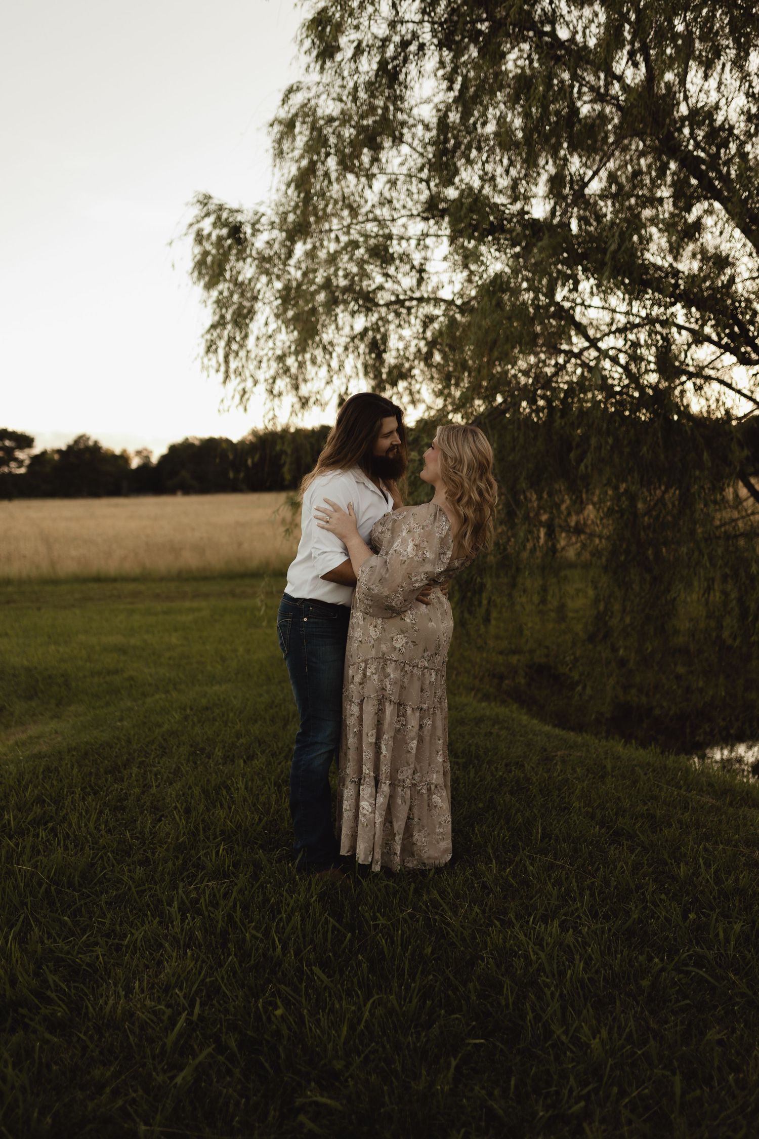 Couple embracing at golden hour near a willow tree by a pond in a lush green field, woman in floral dress.
