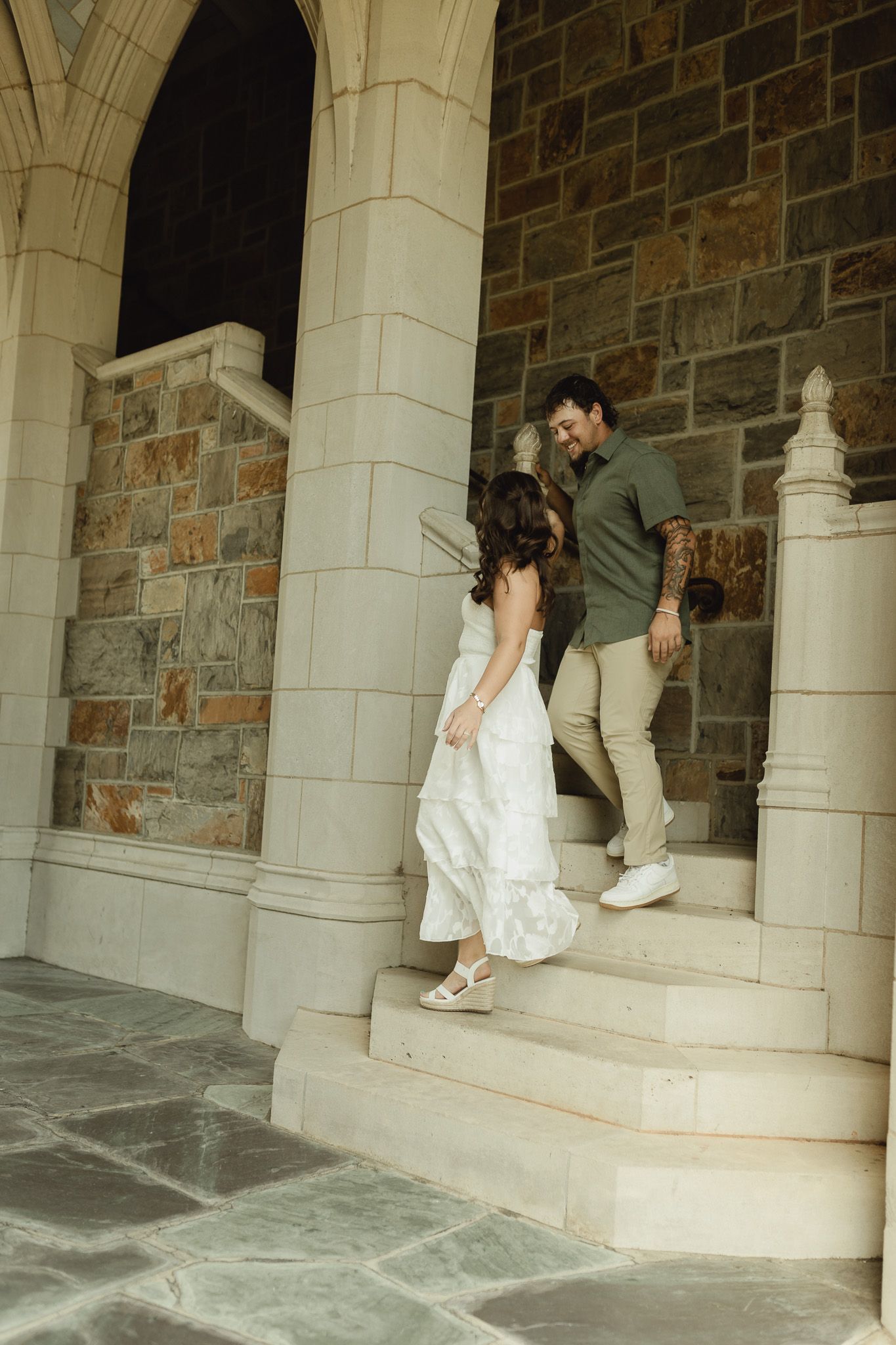 Couple in wedding attire walking up stone church steps, bride in white dress holding groom's hand in green shirt.