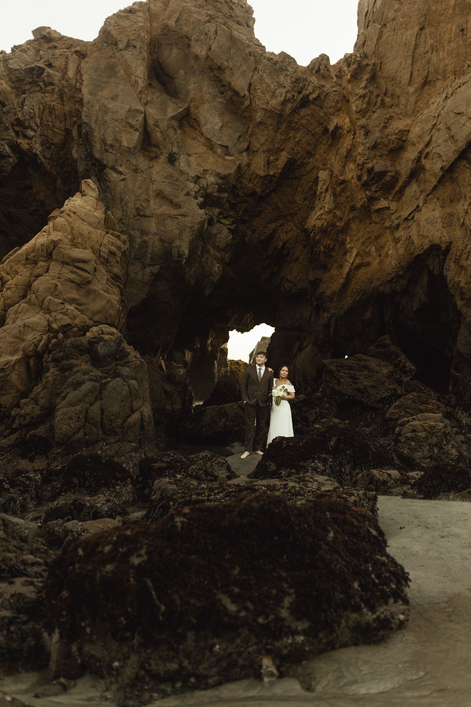 Dramatic coastal wedding scene with couple silhouetted against rocky cliffs and natural arch.