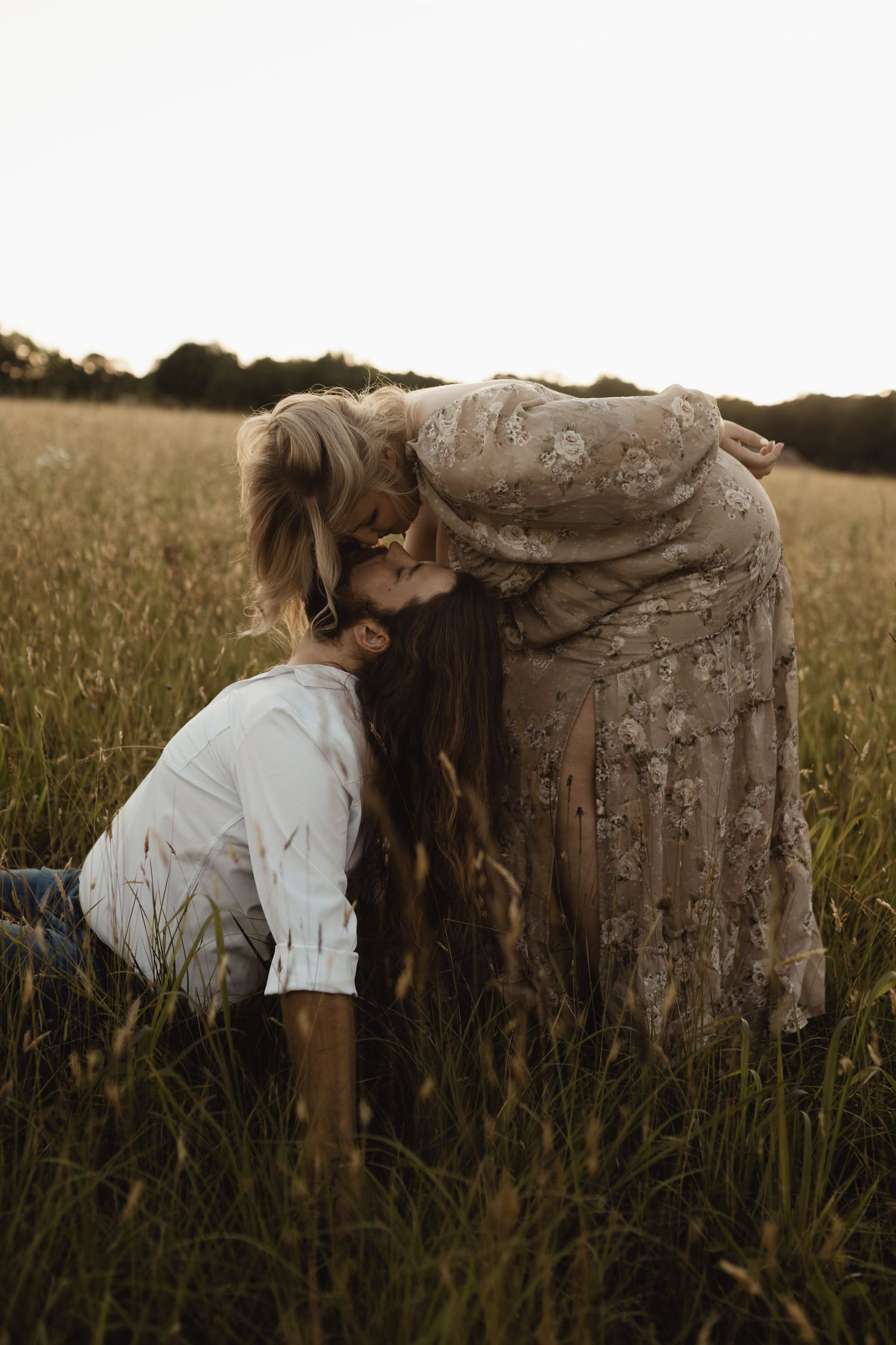 A mother and child share a tender kiss in a golden field at sunset, with the child kneeling in tall grass.