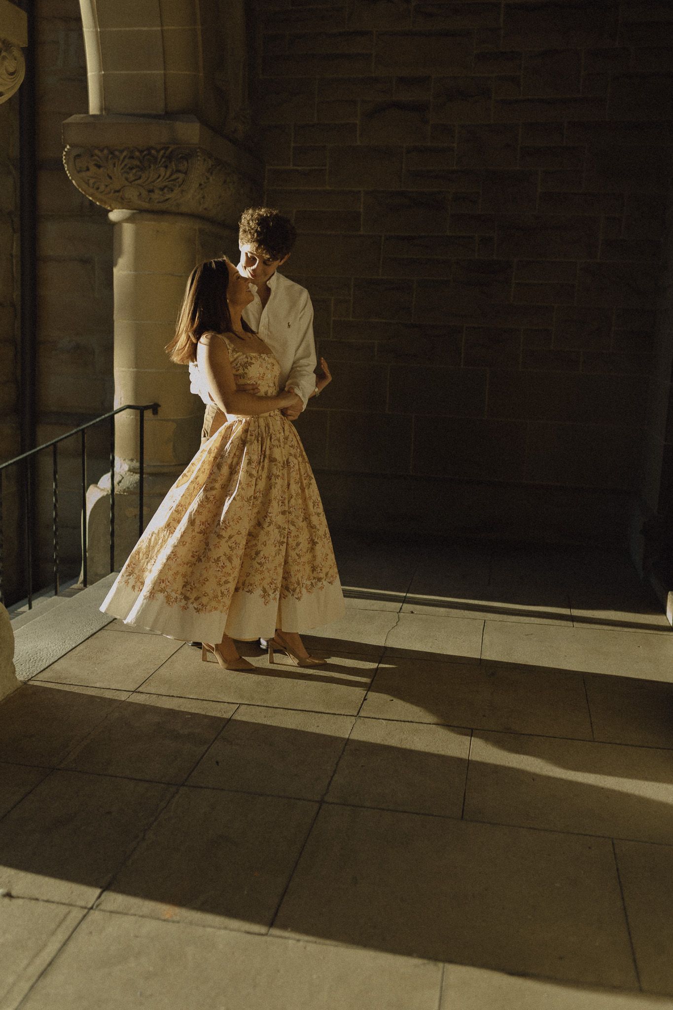 Couple embracing in golden sunlight beside stone columns, woman in elegant floral dress.