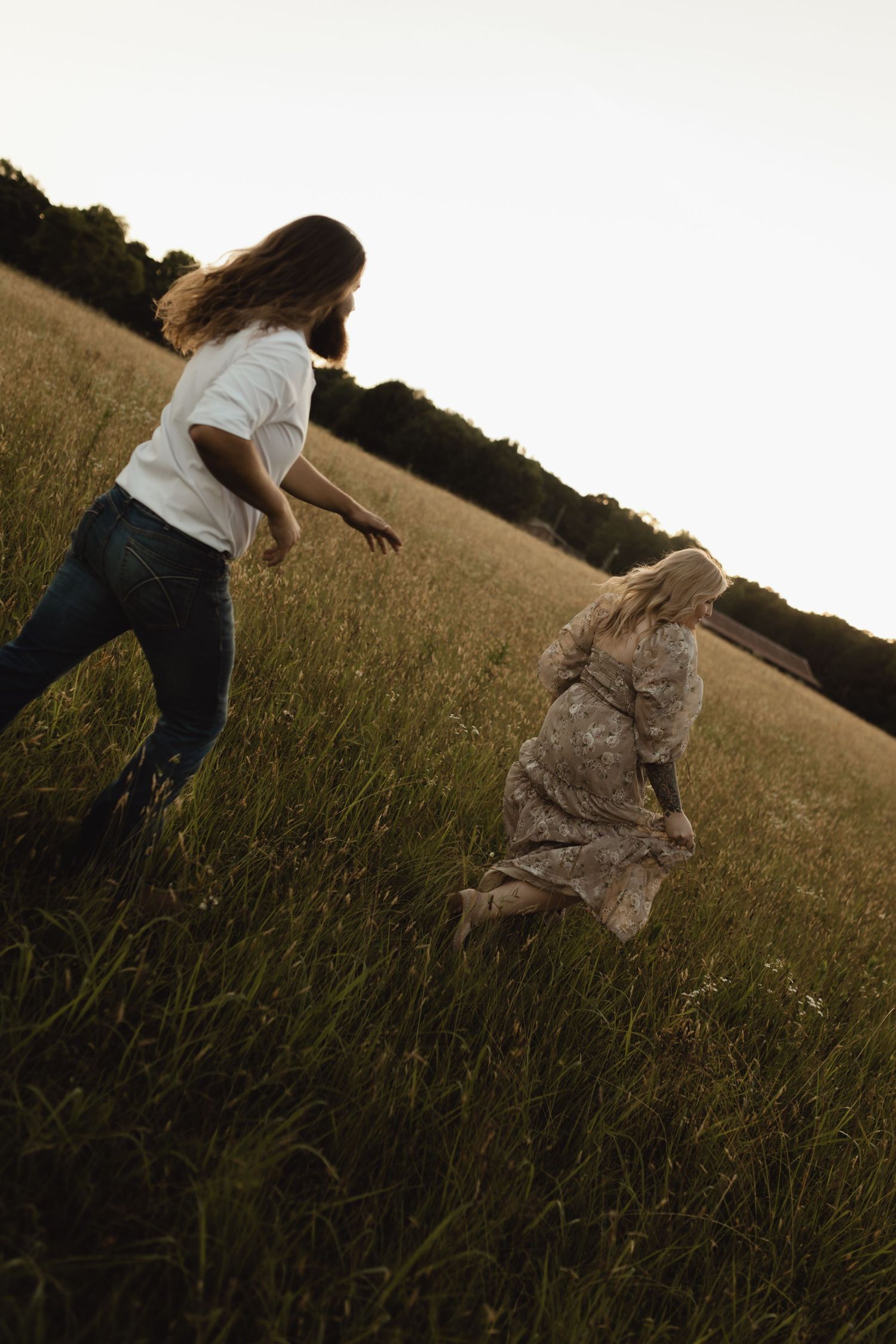 Two women running through a golden grass field at sunset, one in white top and jeans, other in floral dress.