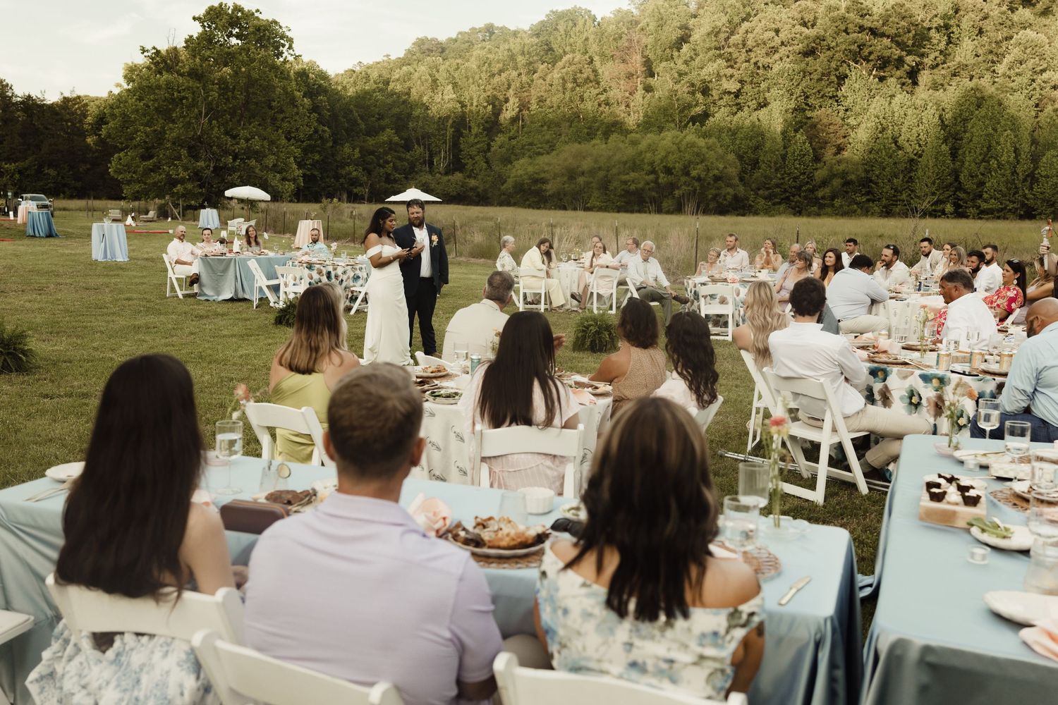 Outdoor wedding reception on a green lawn with guests seated at decorated tables while the couple shares a dance.