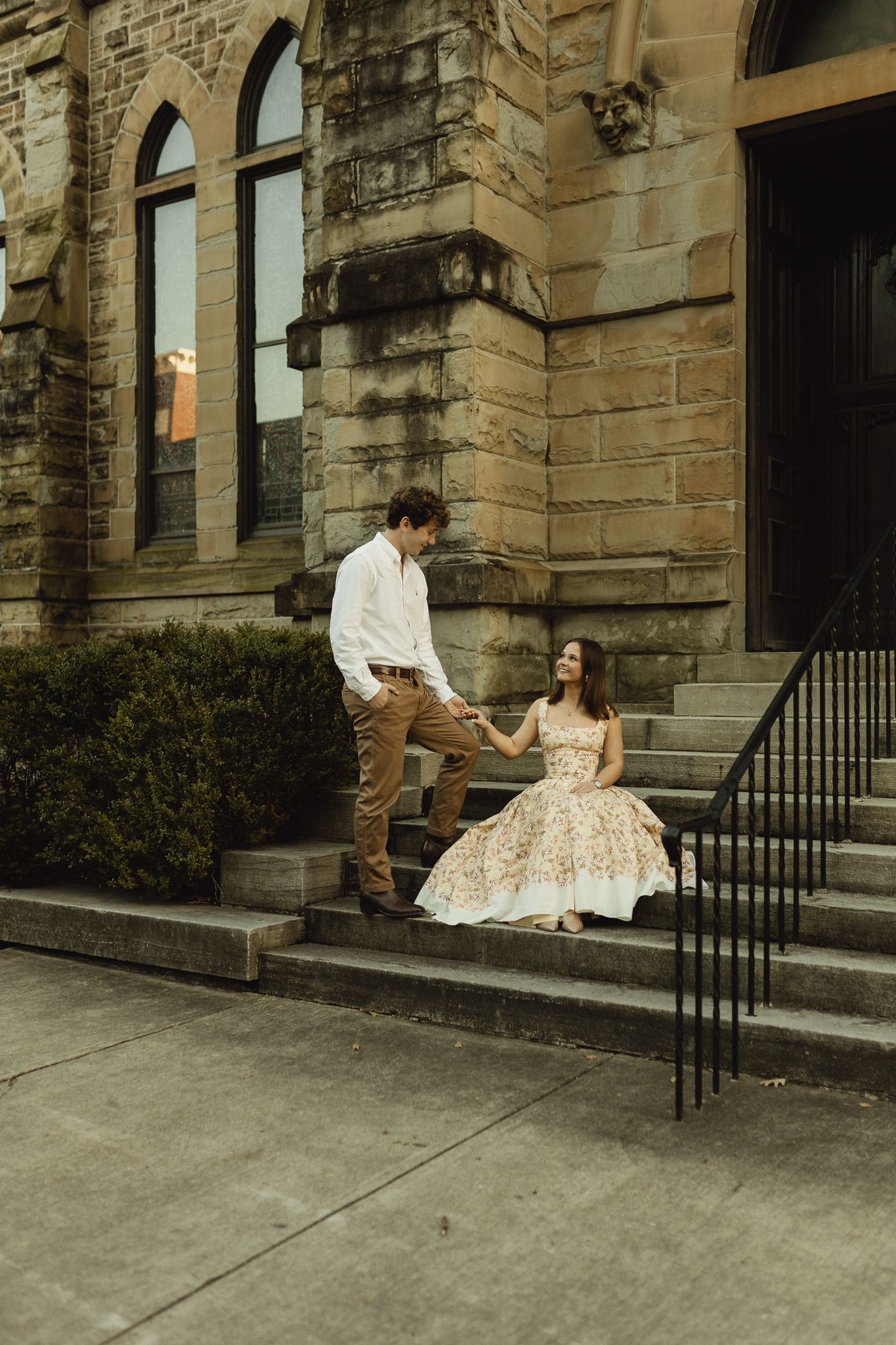 Couple holding hands on stone steps of historic building, woman seated in floral dress.