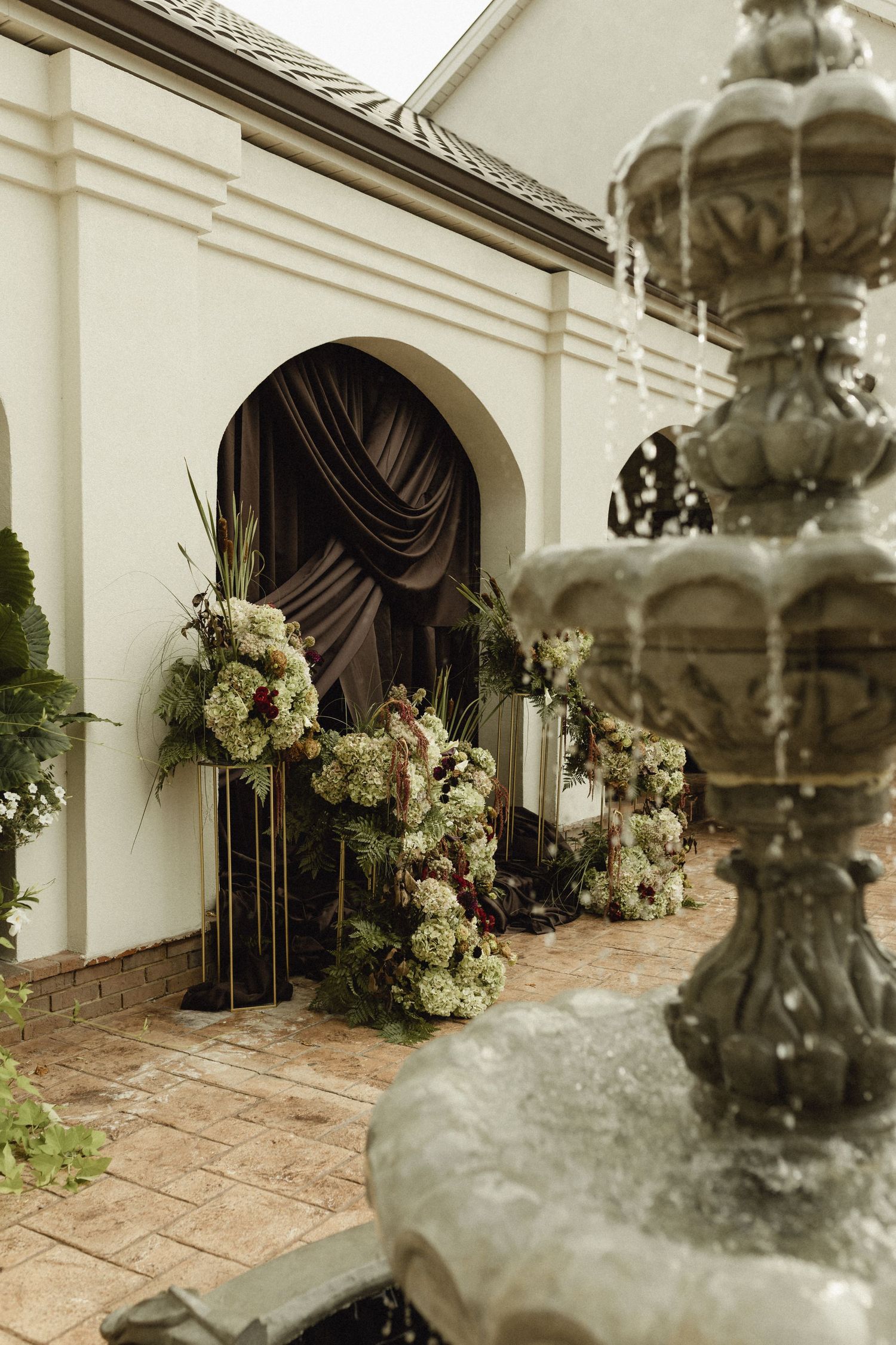 Elegant wedding venue entrance with floral arrangements, dark draping, and a stone fountain in the foreground.