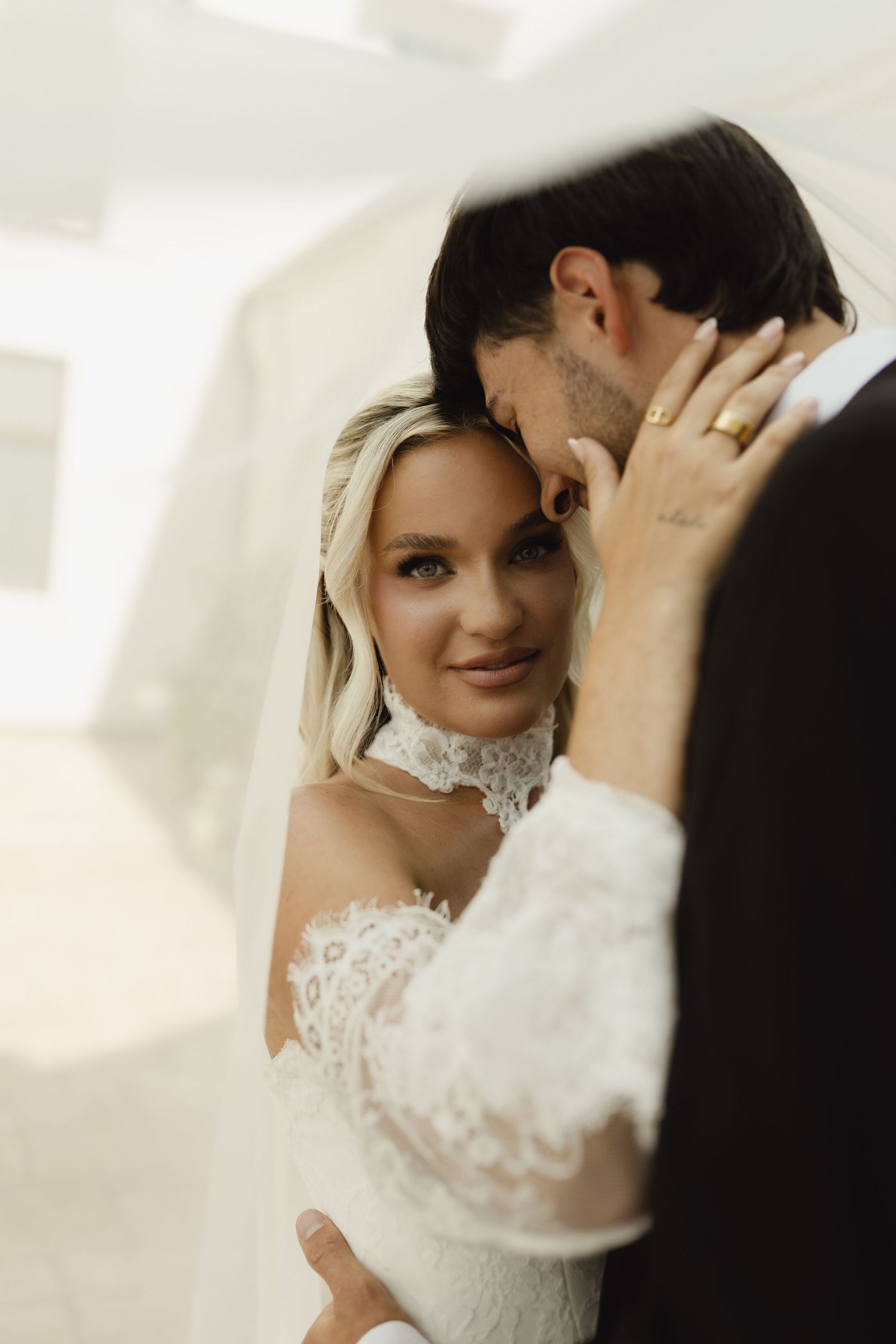 Bride in lace off-shoulder gown and veil embraces groom in black suit during romantic wedding portrait.