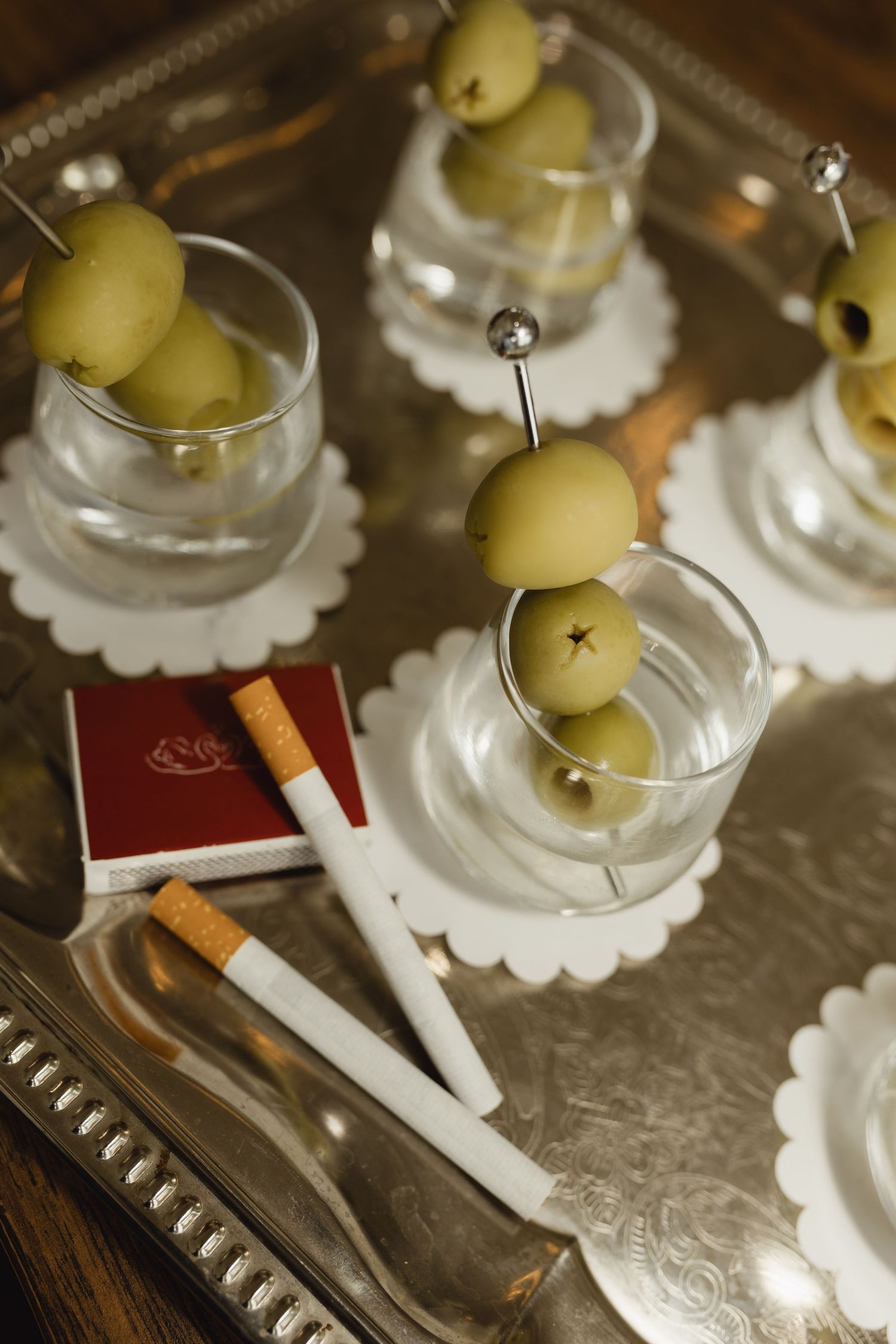 Silver tray with glass cups holding green olives on picks, alongside a red matchbook and cigarettes.
