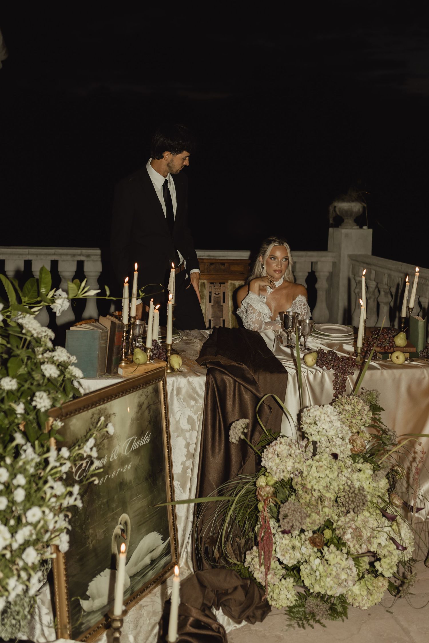 Elegant outdoor wedding reception at night with bride seated at candlelit table adorned with white florals and groom standing beside her.