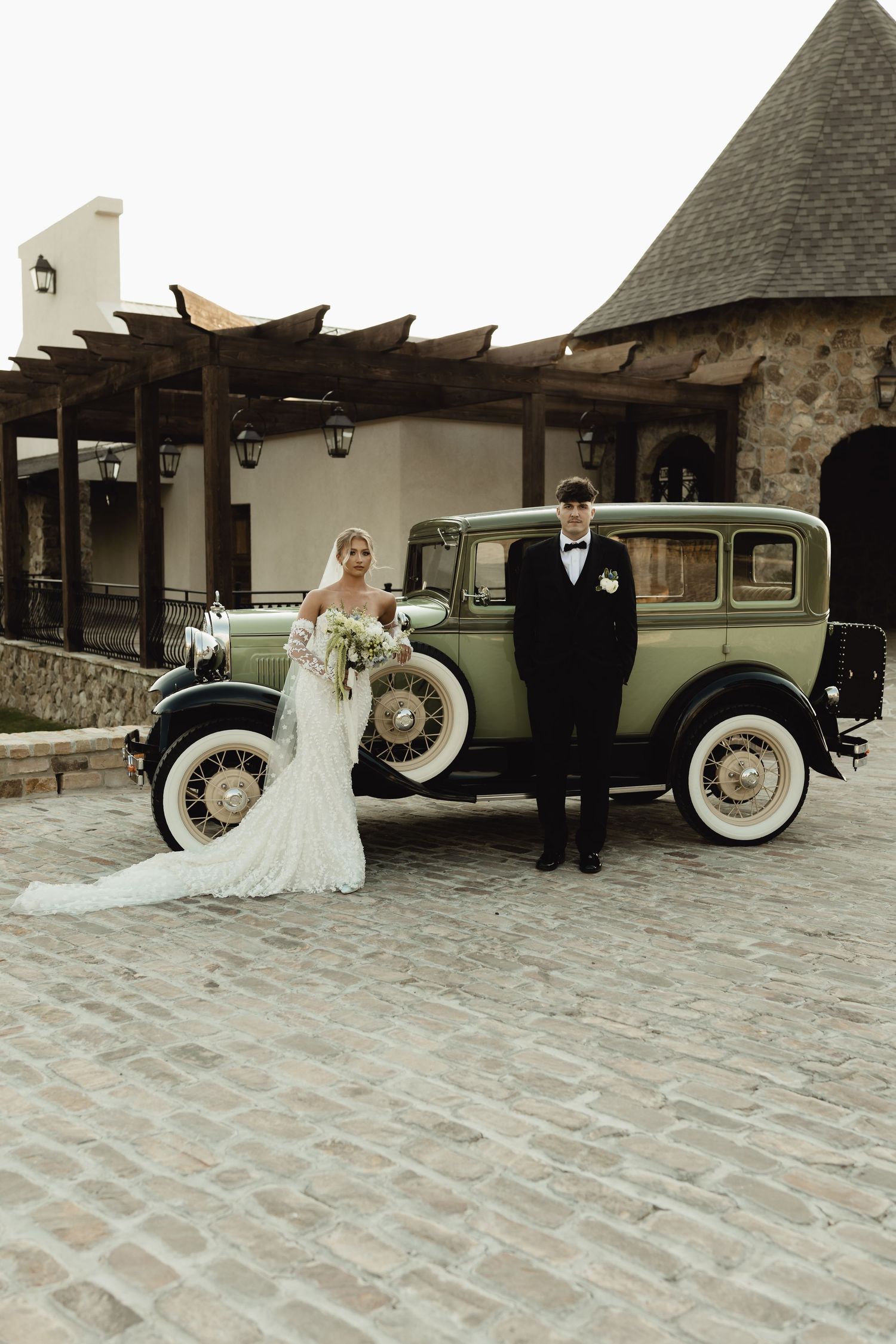 Bride and groom posing beside a vintage green classic car on cobblestone, with a rustic stone building backdrop.