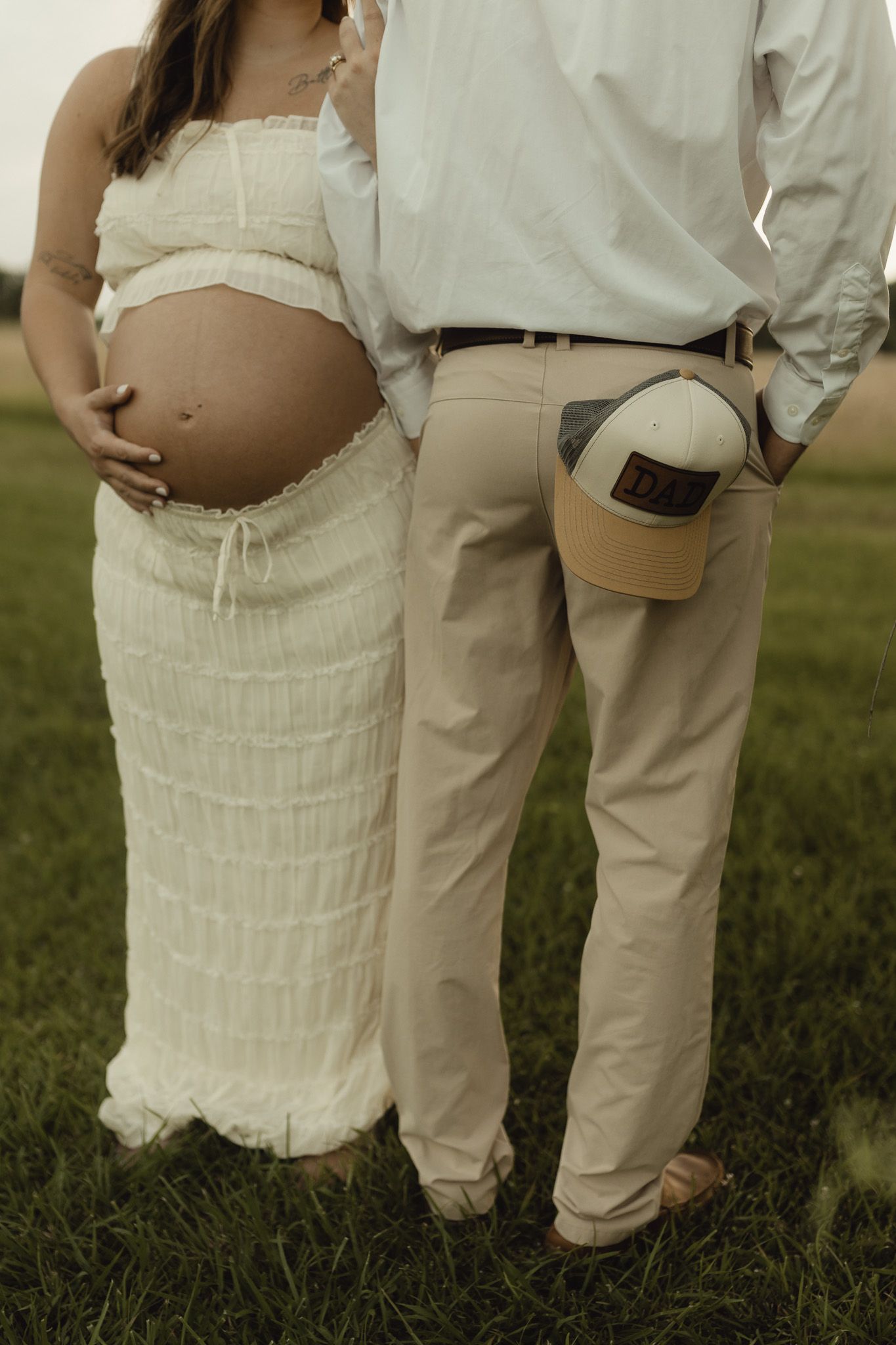 Pregnant woman in white dress stands with partner outdoors, man has a Dad cap tucked in his khaki pants pocket.